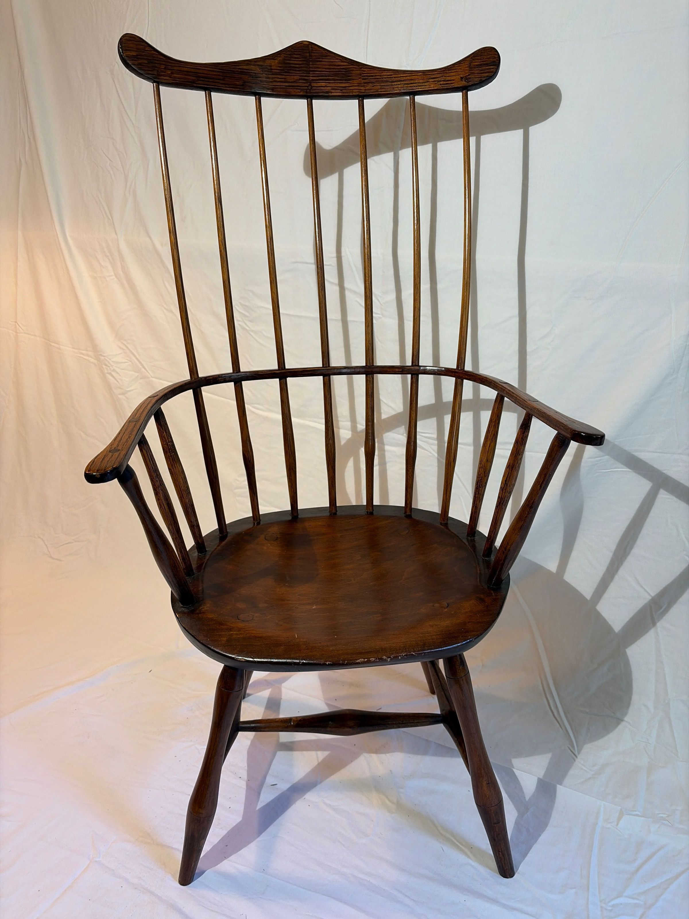 Comb back Windsor wooden chair with a high backrest made of slender vertical spindles, elegantly arching into curved armrests. Its polished seat rests on four sturdy legs connected by horizontal supports, each piece exuding a vintage charm, highlighting the craftsmanship of traditional furniture design.