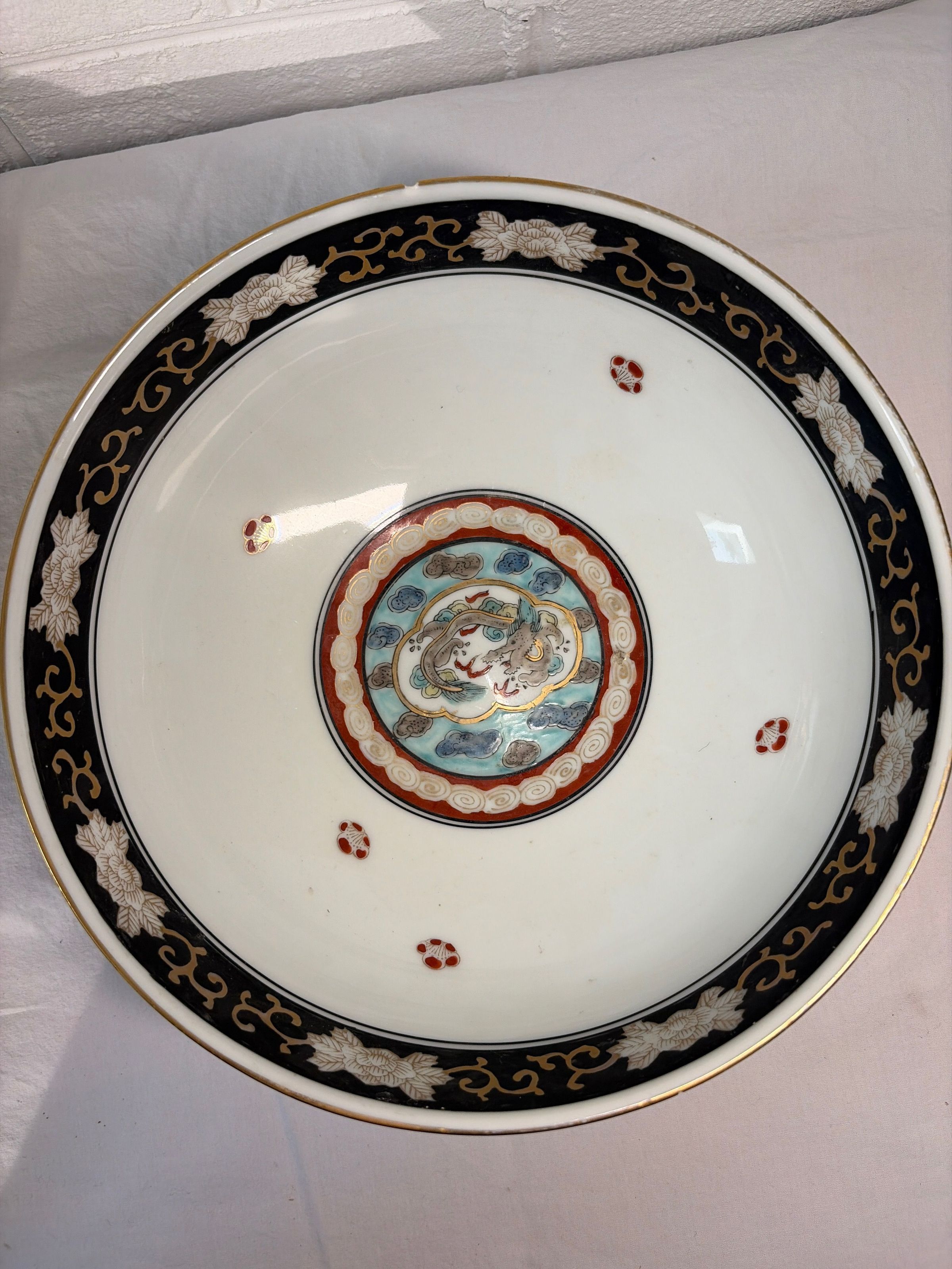 Beautifully crafted porcelain bowl with an intricate design; its interior features a central motif surrounded by concentric circles with blue and red patterns, while the outer edge contains a black band adorned with gold floral designs and white, stylized blossoms, creating a harmonious blend of traditional artistic elements.