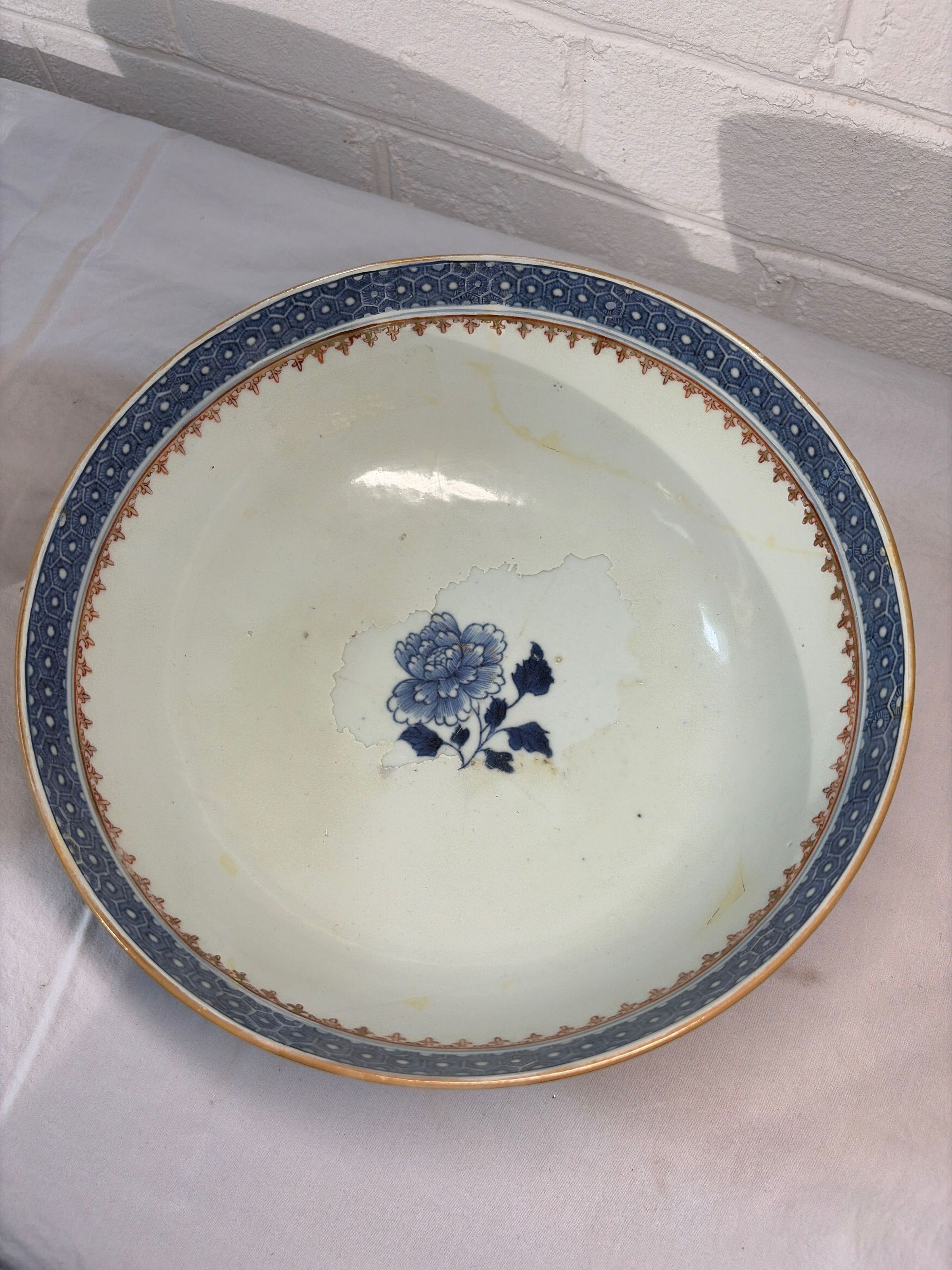 Large porcelain bowl with an intricate blue design on both its rim and center, where a detailed floral motif is featured. The rim of the bowl is decorated with a repeating geometric pattern in shades of blue and gold, adding an elegant and classic touch. The central floral design showcases a finely rendered blue flower with leaves, giving the bowl a traditional and artistic appearance.