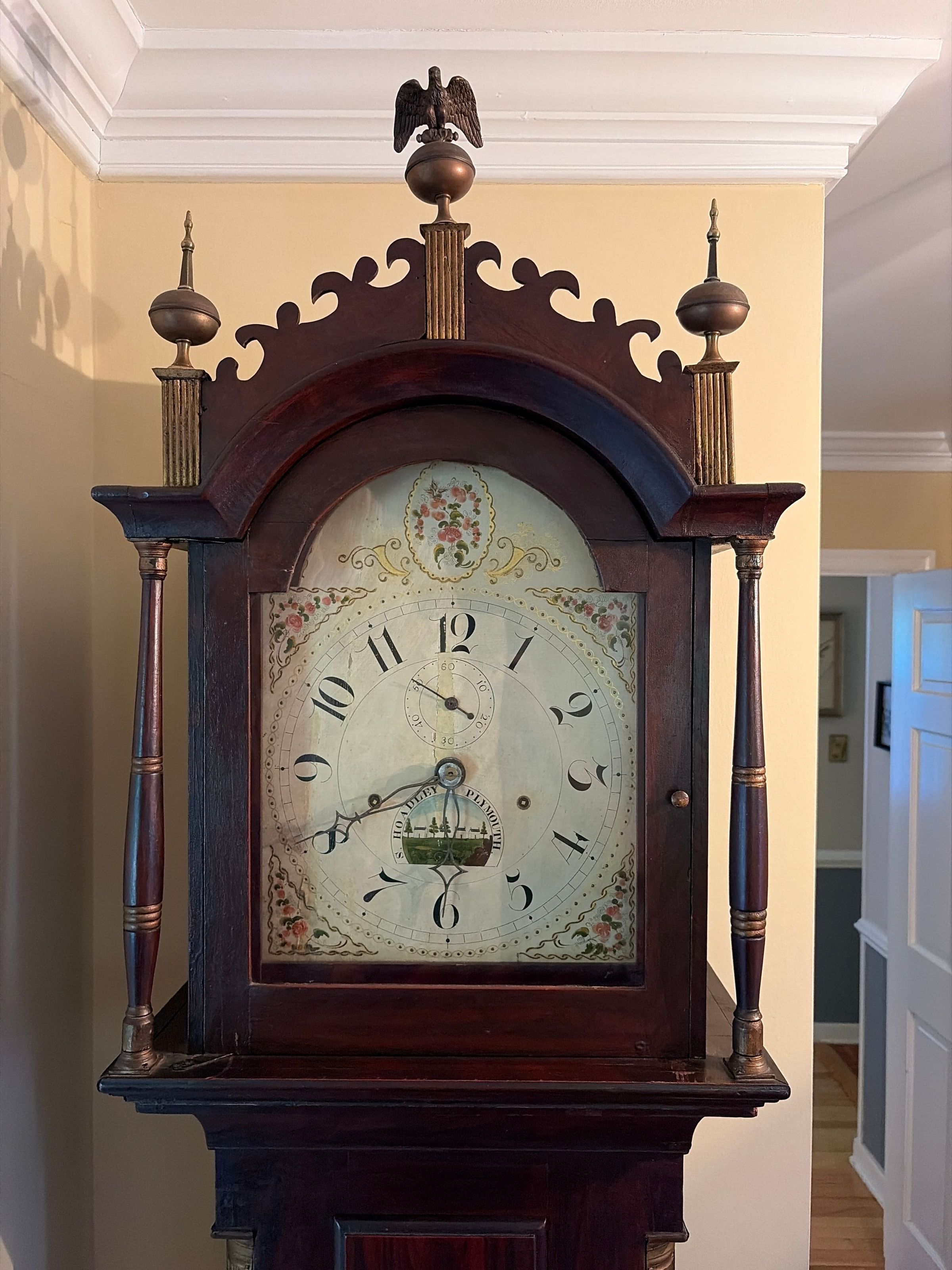 Tall case clock, prominently featuring a wooden housing with decorative elements such as spindles, finials, and an eagle figure perched atop. The clock face displays elegantly crafted numerals with minute and hour hands pointing to the time, adorned with artistic floral motifs and a picturesque landscape scene at the bottom.