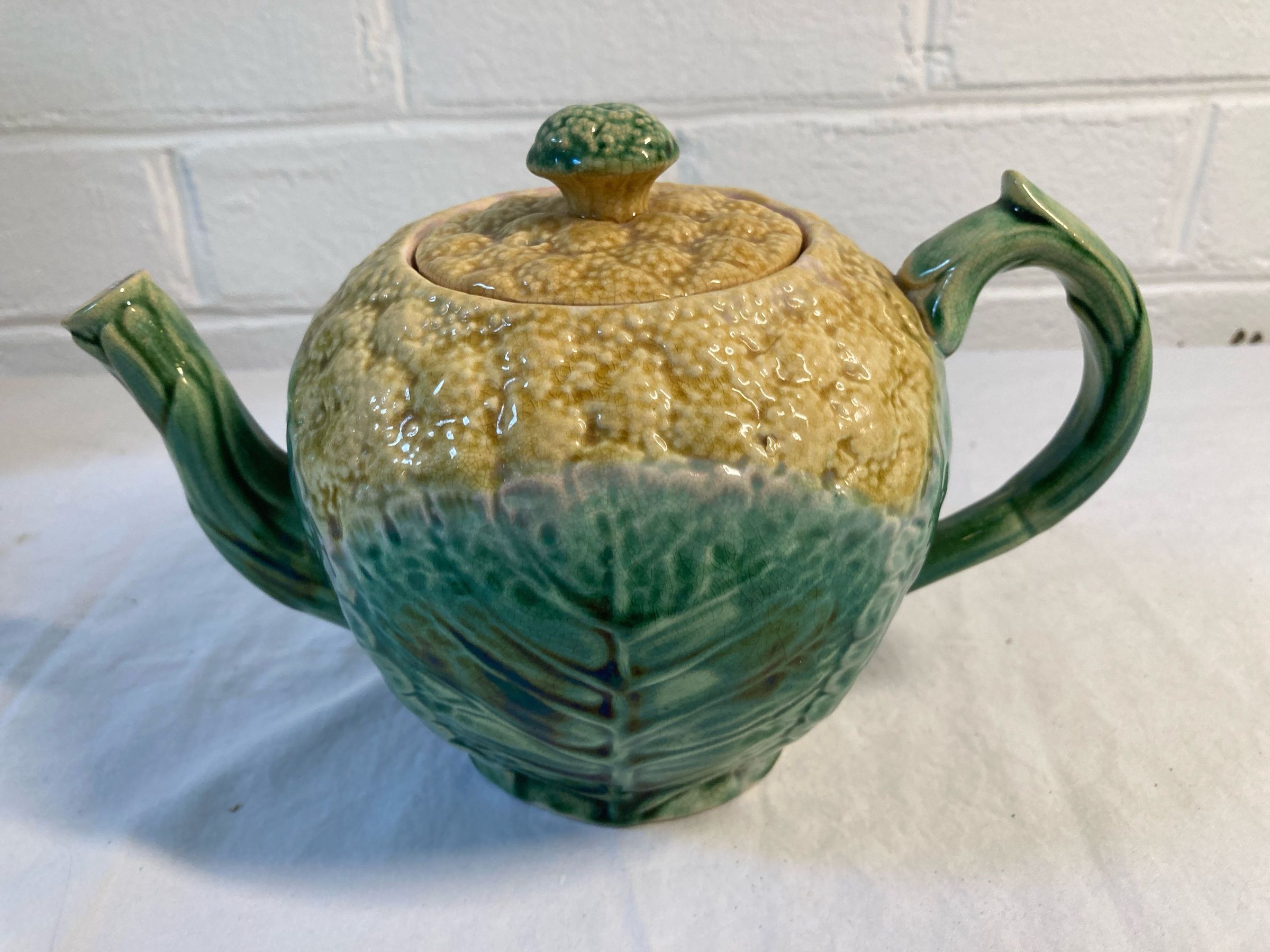 Distinctive Majolica teapot with a textured design resembling a cabbage, featuring a mix of green and yellowish-brown hues that create a natural, earthy aesthetic. The teapot has a round body, a curved spout on one side, and a handle on the opposite side, all crafted with the same organic texture and color scheme. The lid is adorned with a small knob at the top, reflecting the same mottled pattern as the rest of the piece.