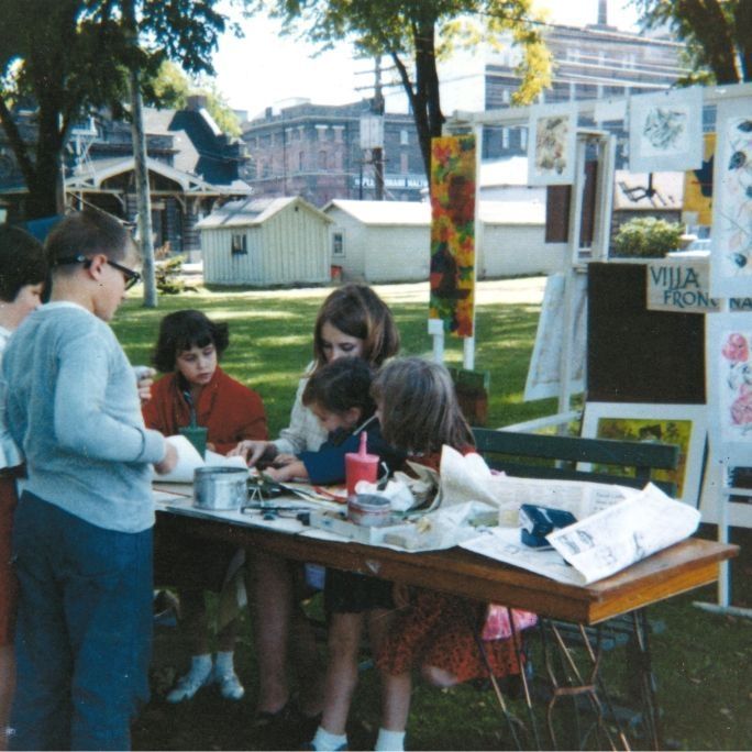 Photograph taken at Red Wing Arts' first Art Festival in 1966: A full-color photograph of a woman helping five children do a craft. The woman and three children are sitting on a bench and two children are standing. They are working on something on a table covered in newspapers. They are outdoors in Levee Park; the weather is sunny and there are numerous pieces of art set up behind them. Text on the back of the photo reads: "Scene from 1966 first Fall Festival in Levee Park."