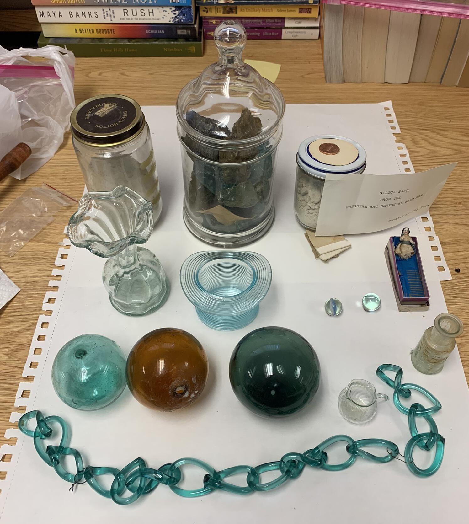 Miscellaneous Blown Glass and Glassmaking Examples: Specifically, the miscellaneous items include from back to front and left to right:
- Three jars of glassmaking sand and slag, including one which is labeled, "Silca sand from the Cheshire and Berkshire sand beds. Donated by Judy Rowe."
- A blown glass vase, pressed glass dish, two blown glass marbles, and a toy or souvenir glass doll.
- Three blown glass balls, a toy blown glass pitcher, and the Dr. W. Evans bottle. Town Historian Judy Rowe suggested that the Glass balls were used for knitting during her "Glass Lake Industry" program presentation. Judy also suggests that the marbles, tiny pitcher, and other blown glass items may have been "whimsical" creations that glassmakers typically made for fun toward the end of their production shifts. Refer to the DVDs of program presentations entry in this folder for additional information.
- A pulled glass chain.