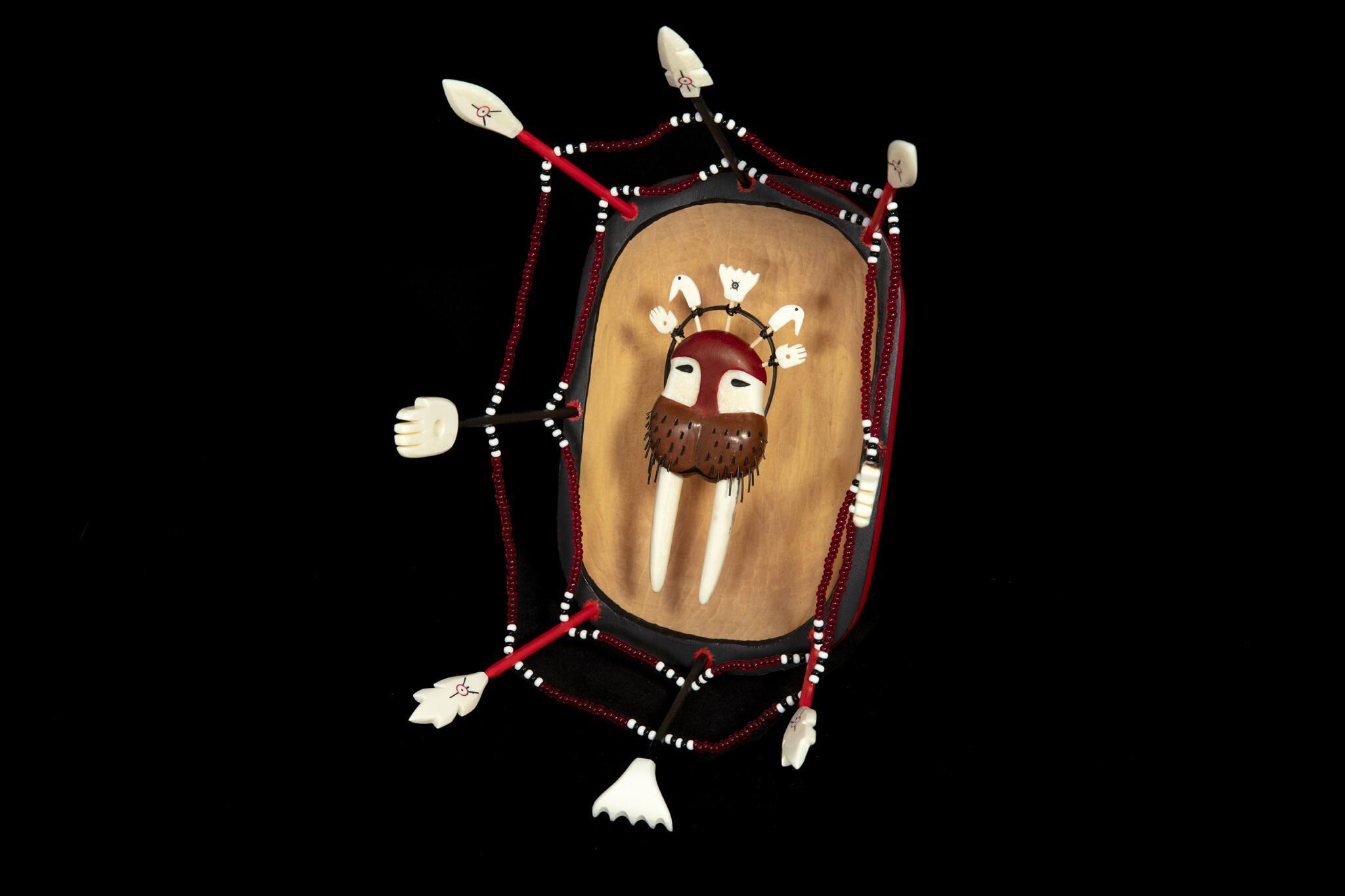 “This mask is made to represent the transformation of the walrus’ spirit from the spirit world into our world. The face of the mask represents the walrus and human spirit. You can see the human spirit in the shape of the eyes. The hoop represents the emergence of the walrus’ face from the spiritual realm. The hands with the holes in the middle represent the passageways where the walruses come through for abundance. The harpoon on top represents the hunters who will be hunting the walrus. You can see how it appears the walruses are going towards the harpoon. The circle and dot symbol seen on the walrus and harpoon pieces is the ‘eye of awareness’, showing respect to the creator of the universe, and also representing the richness of Yup’ik culture.” – Mike Wassilie