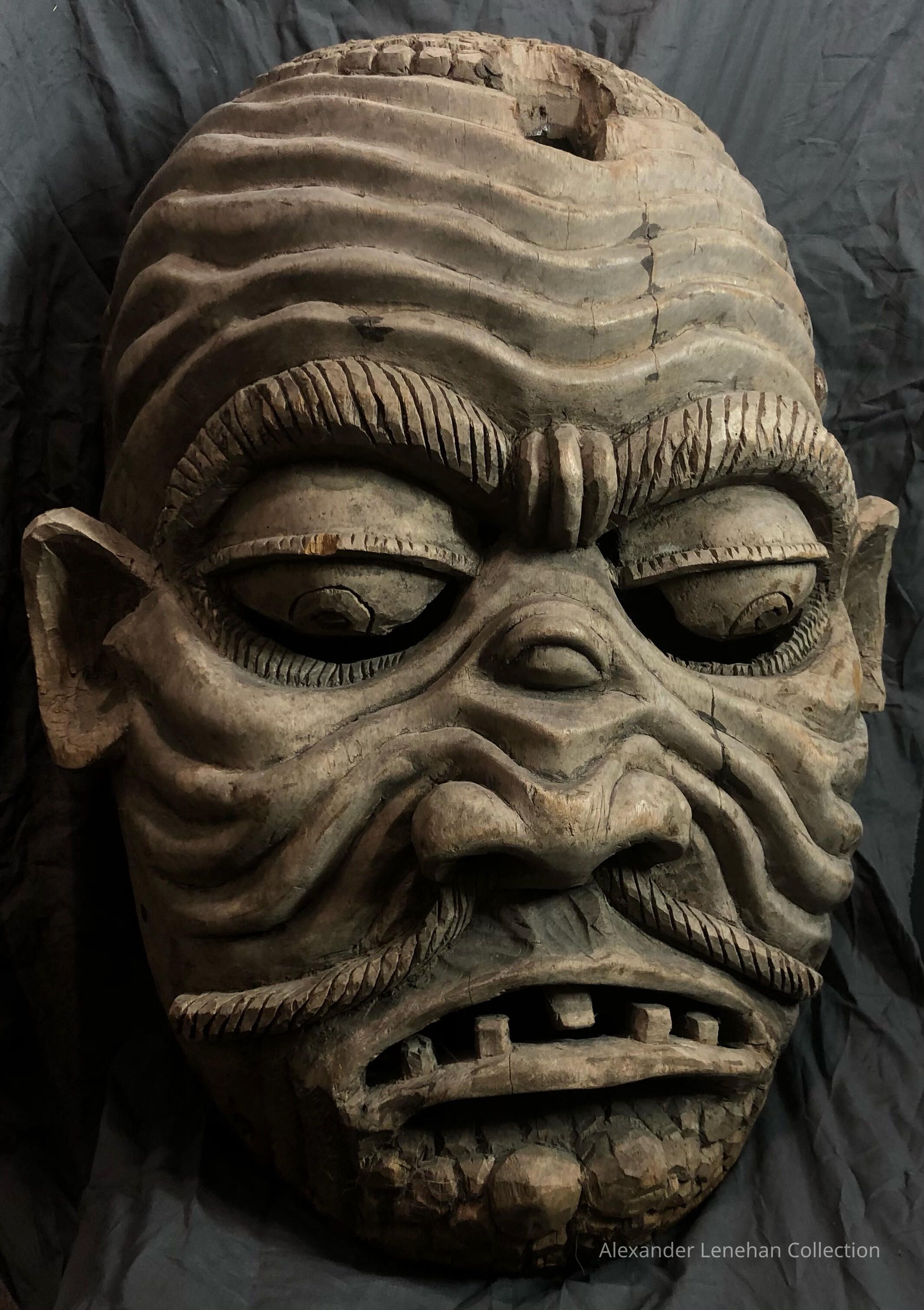 The image displays an intricately carved wooden mask featuring a highly expressive face with detailed, deeply furrowed eyebrows, piercing eyes, and an open-mouthed scowl. The textures and contours of the wood, including apparent cracks and age marks, suggest historical value and craftsmanship, enhancing the mask's formidable and intense appearance. The background of the image is a simple, dark fabric that contrasts with the lighter tones of the aged wood, focusing attention on the mask's dramatic expression and artistic detail.