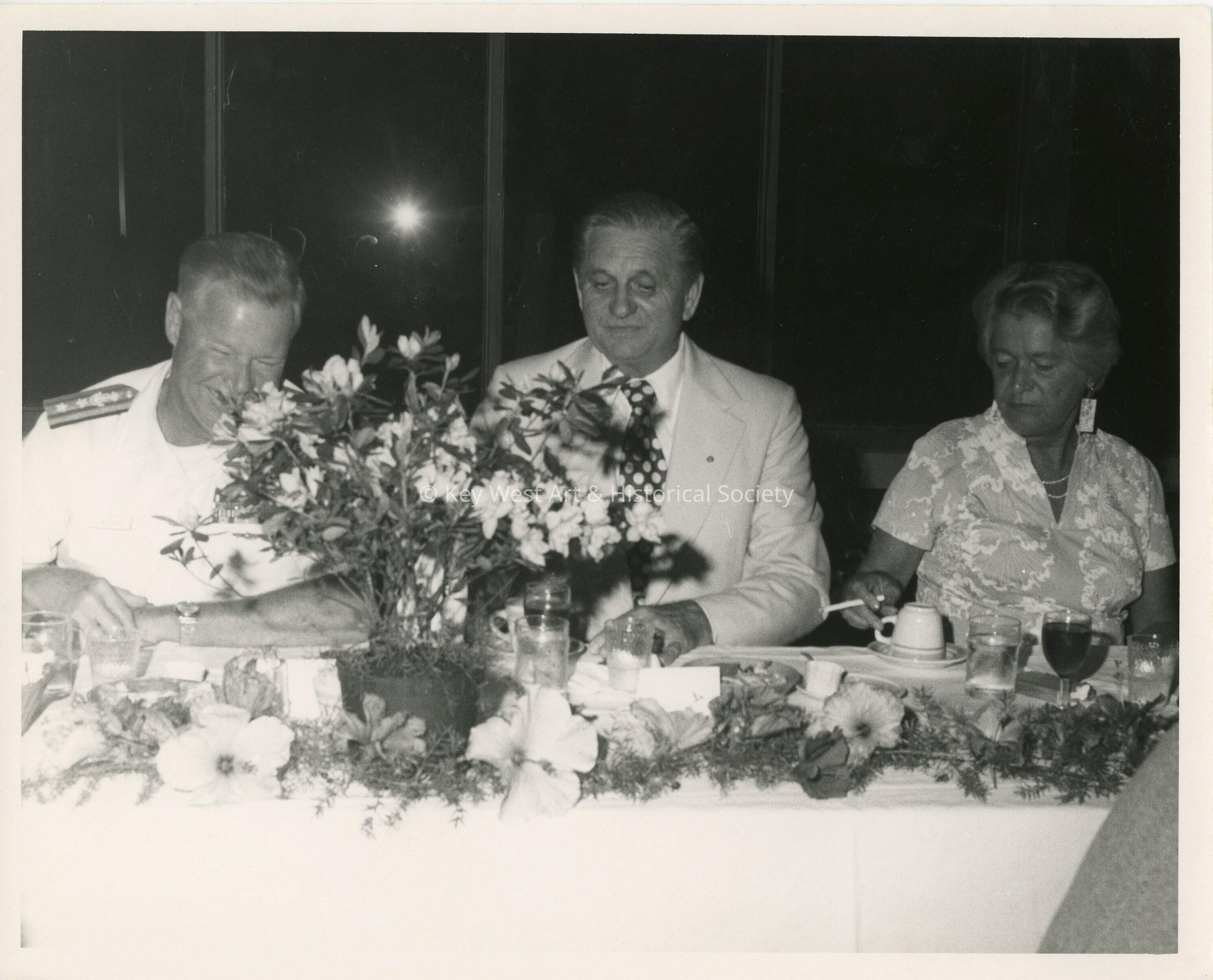 3 Unknown people sitting at a table at a party, one is in uniform