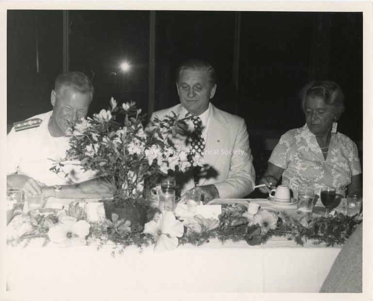 3 Unknown people sitting at a table at a party, one is in uniform