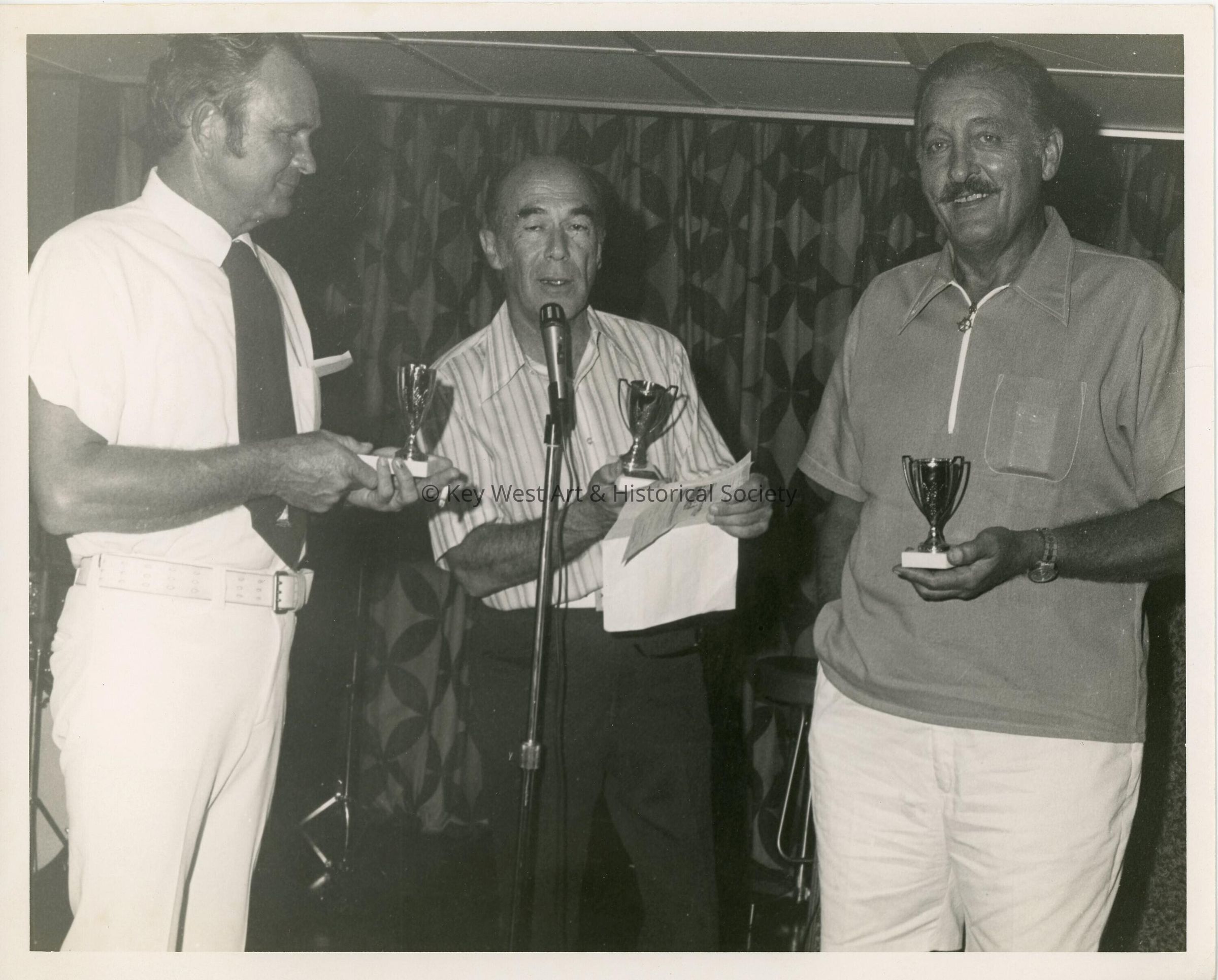 3 Unknown men standing at a microphone each holding a  trophy