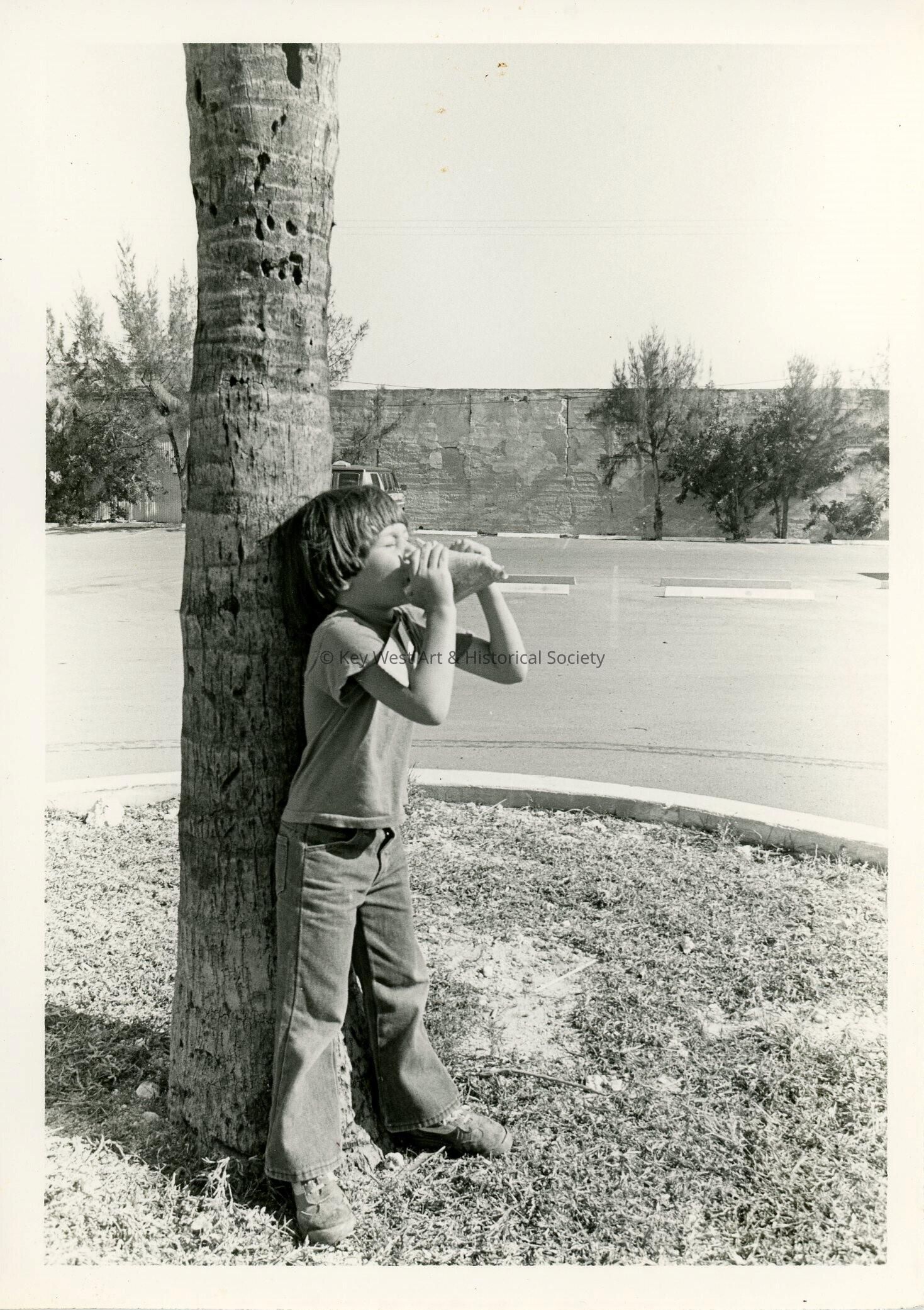 A child leaning against a tree blowing a conch