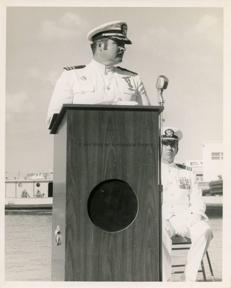 2 Men in uniform, one sitting and one standing at the podium