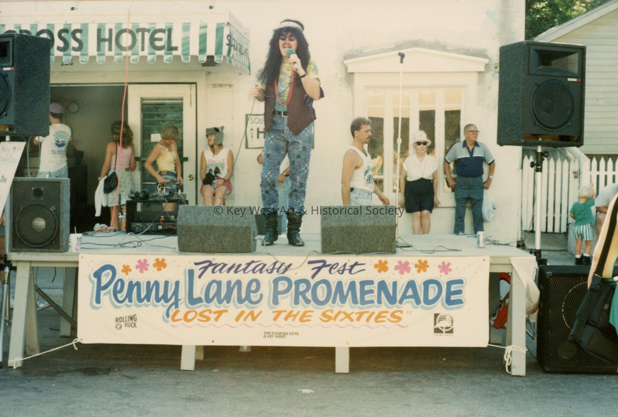 An unknown person dressed up on a stage. Sign reads Penny Lane Promenade.