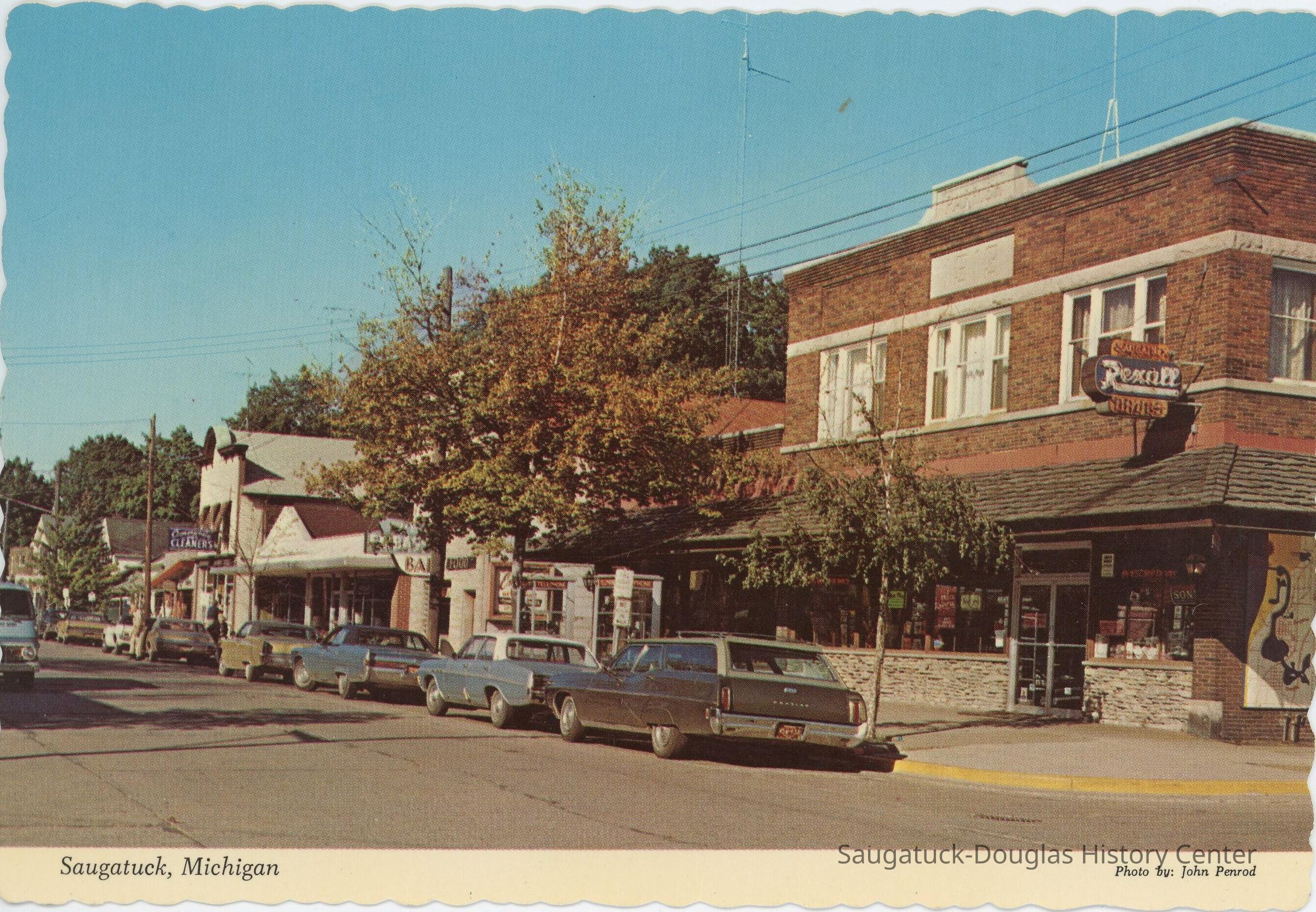 Street view of Saugatuck Postcard; Several cars line a street in Saugatuck with shops in the background.