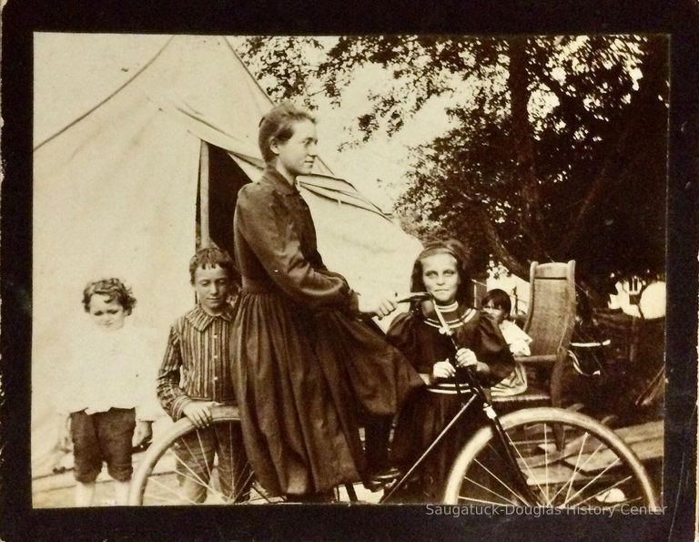          Melcher Family at Pier Cove, Michigan; Woman on bicycle in front of tent, with four children posing toward the camera.
   