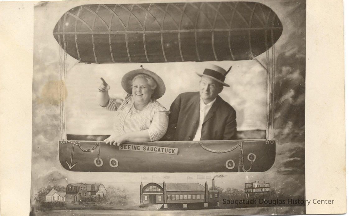          04.BalloonEyeViewofTown.SUPER!!!.jpg 100KB; A man and a woman in a photo background that look like they are in balloon floating over Saugatuck landmarks the Coral Gables (then Hotel Saugatuck), Big Pavilion and Hotel Butler. A sign by her arm reads 