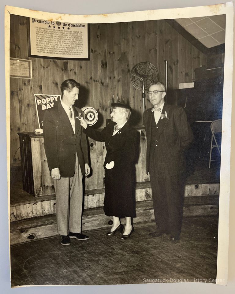          Memorial Day 1954; Lillian Morris pins poppy on lapel of Mr. Sholders[?] the president to Douglas village with Mr. George Wright, president to Saugatuck Village. Location: American Legion post 137 hall.
   