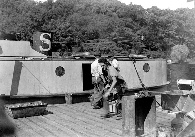 The tug Andrew was owned by Frank Sewers. It docked at Water Street, across the river from the Saugatuck-Douglas History Museum. A typical tug of the era, Andrew was enclosed to protect fishermen from the weather and to provide an enclosed workspace. Note buoy on roof of tug. Man dragging net boxes is Ev Van Plew