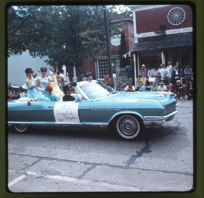 Page D 18. Miss Saugatuck Douglas 1968, Parade queens waving from back of convertible, on Butler Street in front of Bay Building.; Bay Building 110 Butler Street and Country Store Antiques at 120 Butler Street
