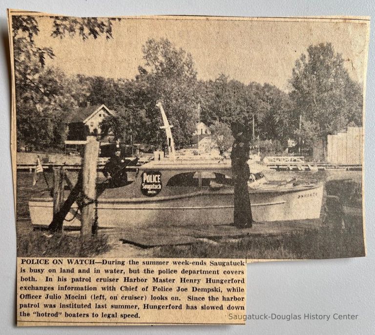          Newspaper clipping from unknown publication; POLICE ON WATCH- During the summer week-ends Saugatuck is busy on land and in water, but the police department covers both. In his patrol cruiser Harbor Master Henry Hungerford exchanges information with Chief of Police Joe Dempski, while Officer Julio Mocini (left, on cruiser) looks on. Since the harbor patrol was instituted last summer, Hungerlord has slowed down the 