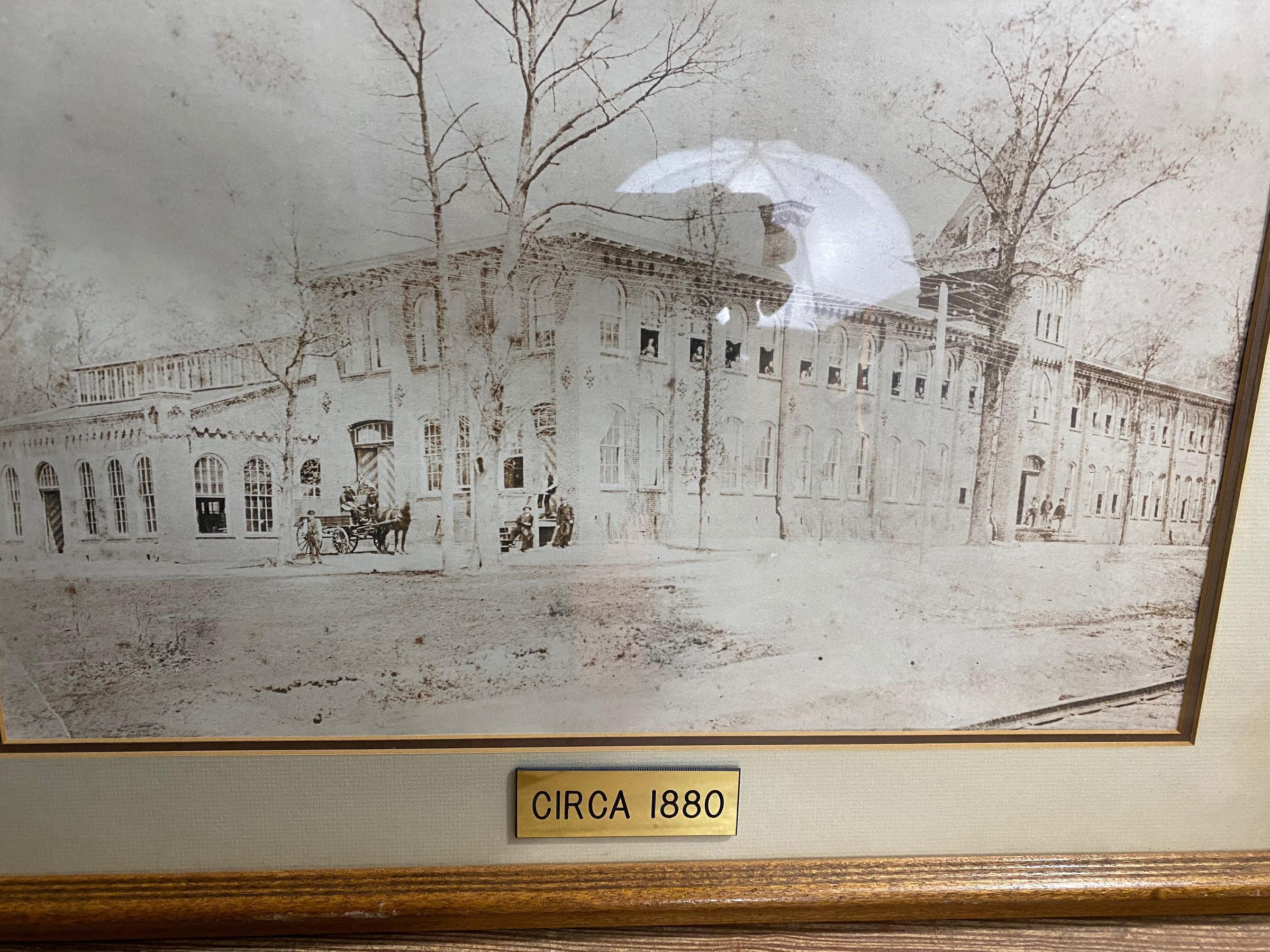 Black and white photograph of Springfield Worsted Mill (site of Ocean Spray Company); in brown wood frame with off-white mat; gold plaque on bottom center of mat reads "CIRCA 1880": A horse-drawn carriage is positioned to the left side of the building with three men on the carriage, and one men standing in front. Three other men are positioned closer to the building to the right of the carriage: one standing, two leaning/sitting on the railing of the stairs from the door. Three more men stand at what appear to be the main entrance, on the steps in front of the building, on the right side of the photo. At least 11 people appear to be looking out of open windows on the second floor of the building.