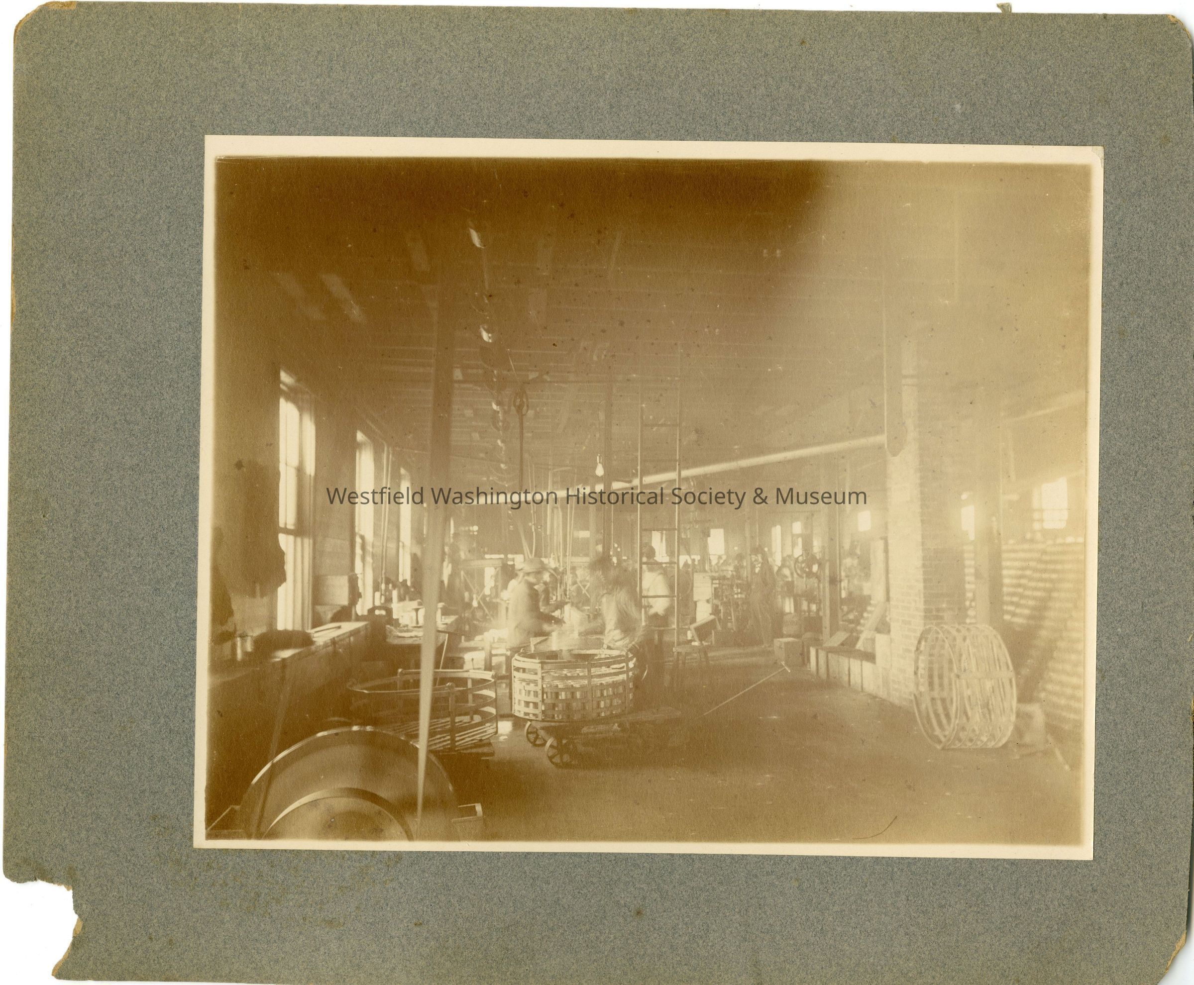 This sepia-toned photograph captures an industrial scene inside a large factory, where the spacious room is filled with various machinery and equipment, conveying the bustling atmosphere of early 20th-century manufacturing. In this expansive setting, light streams through multiple rectangular windows along the left wall, illuminating the interior and casting shadows on the wooden floors. Central to the image are several workers, indistinct but visibly occupied, engaged in operating and monitoring the machinery that lines both sides of the room.