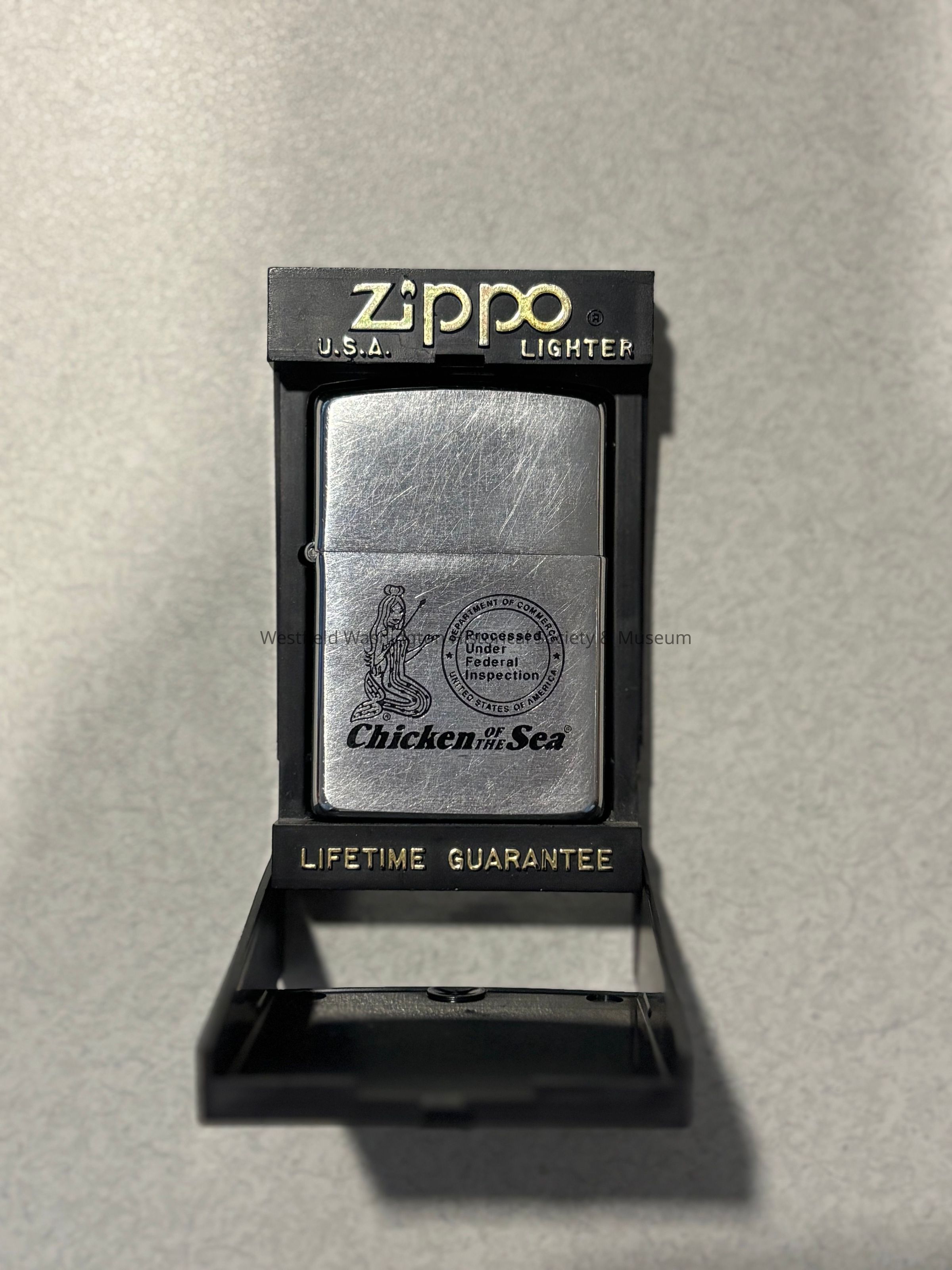 The image features a silver Zippo lighter displayed in a black stand with the word "Zippo" inscribed at the top, alongside "U.S.A. Lighter," and below the lighter, it reads "Lifetime Guarantee" in gold letters, while the lighter itself bears a "Chicken of the Sea" logo and what appears to be a mermaid design along with a seal indicating "Processed Under Federal Inspection,"