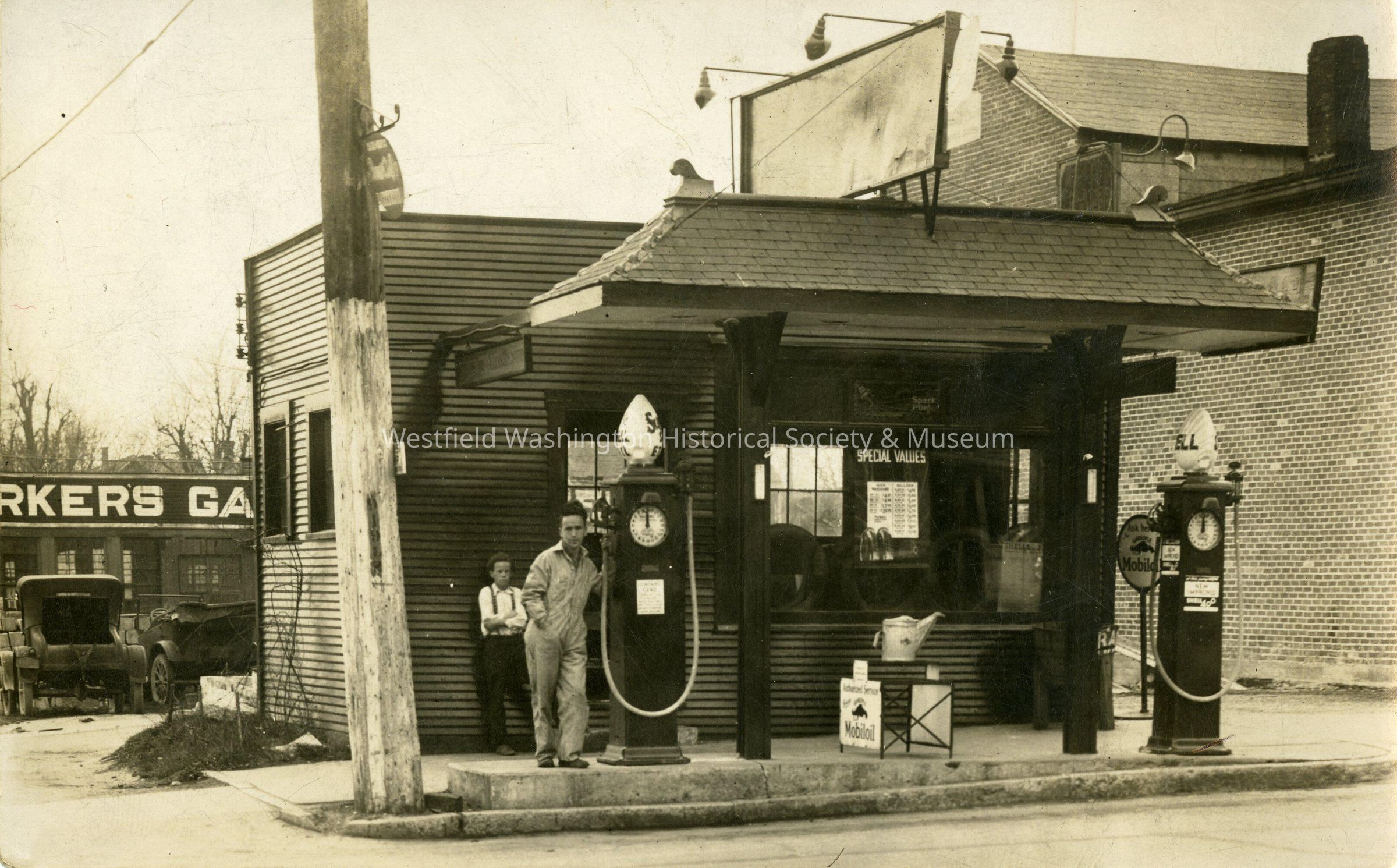 Don and Bob Barker at the Barker Filling Station, South Union Street, Westfield, Indiana: The black-and-white photograph depicts an old gas station, featuring a small, wood-clad building adorned with signage such as "Workers Gas" and "Special Values." Classic fuel pumps line the forecourt, where a man in a work outfit stands by one of the pumps. Off to the side, a young man leans against the building, observing the surroundings. The street scene is completed with details like an early model car parked nearby and various advertising signs.