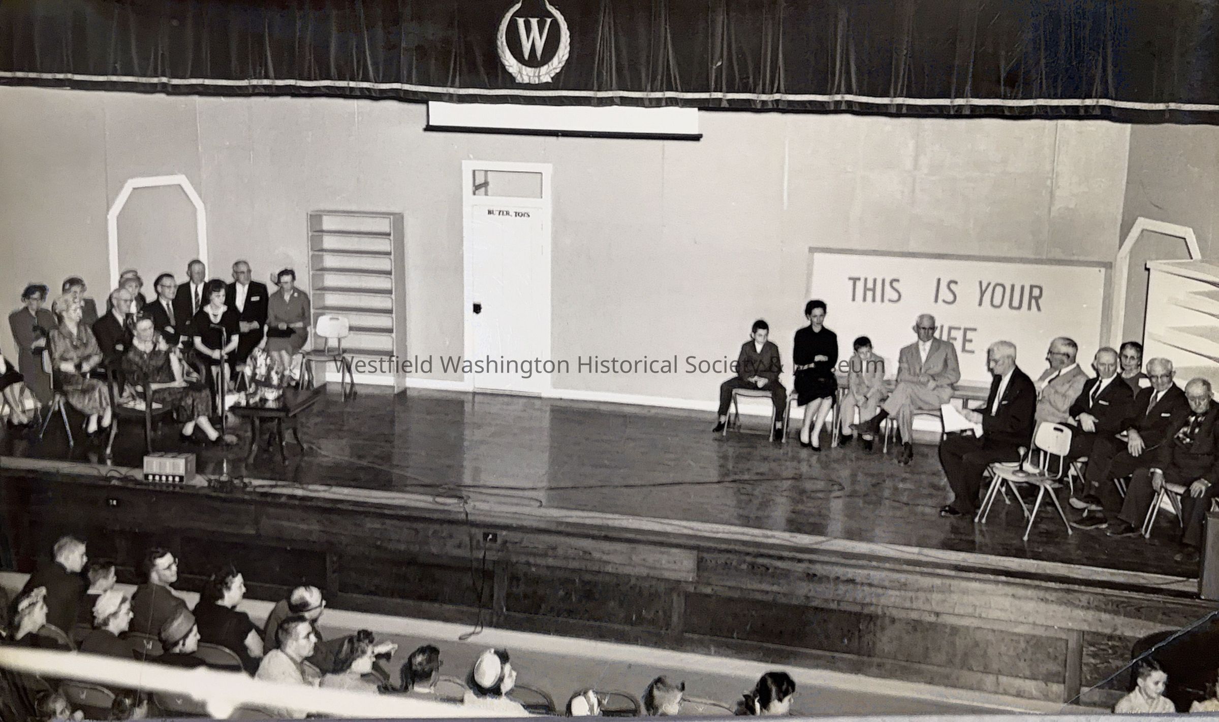 The black and white photo depicts a stage set up reminiscent of a mid-20th-century theater production, featuring a group of men and women seated in two separate sections, with a visible audience in the foreground. On one side of the stage, a group of well-dressed women and men sit closely together, while on the other side, individuals are more spaced out, flanking a large sign that reads "THIS IS YOUR LIFE." The stage backdrop features a simple design with a door and empty shelves. A microphone is seen on each side of the stage, one for each section.