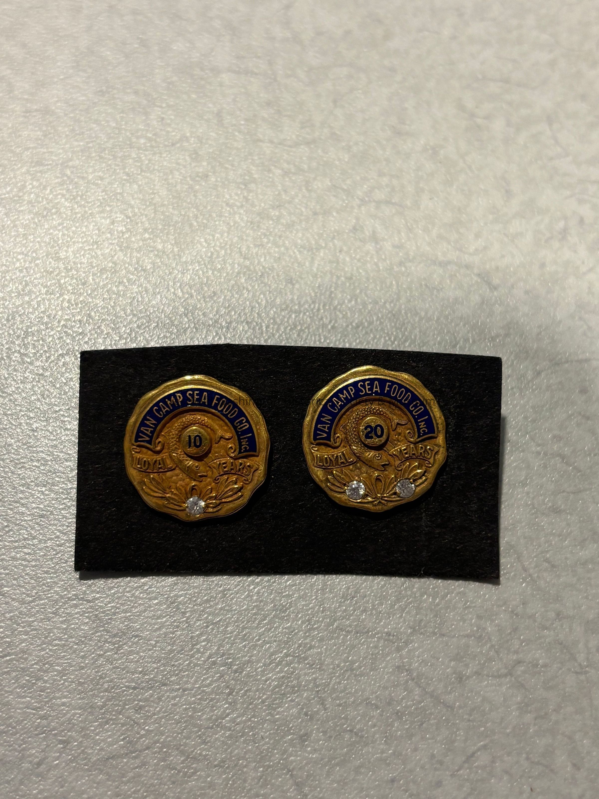 The image displays two gold-tone pins placed on a black rectangular background, each with intricate detailing and a small gem at the bottom. The pins commemorate years of service, with one showing "10 Loyal Years" and the other "20 Loyal Years," both marked with the name "Van Camp Sea Food Co Inc." The design includes a curved blue banner and ornate floral elements at the base, emphasizing the significance and appreciation of dedication over time.