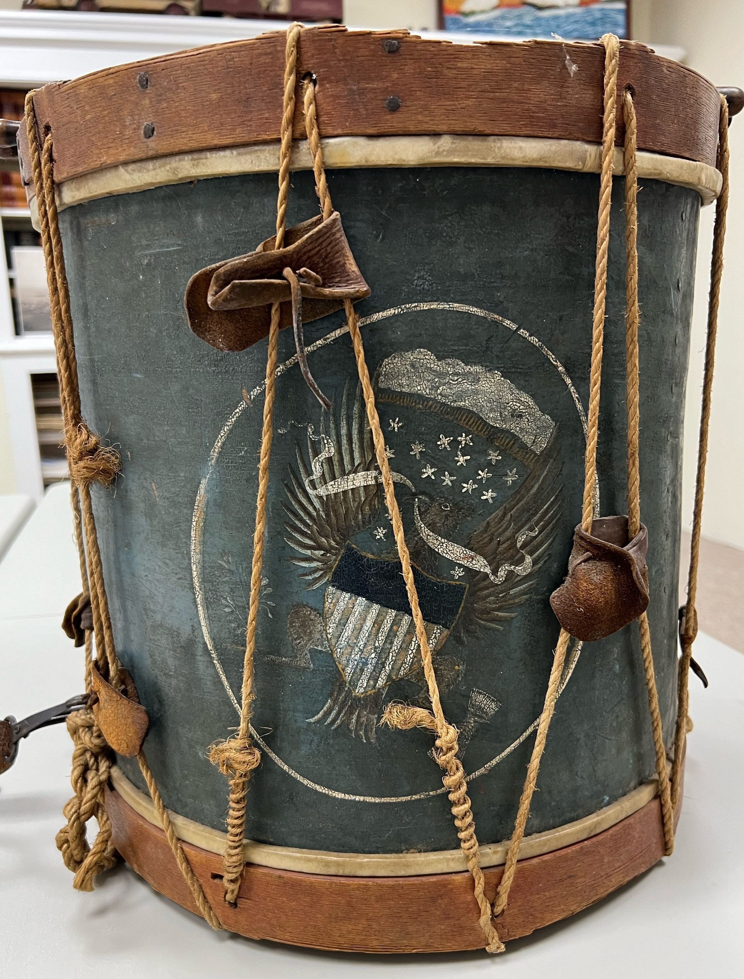 The image displays an old, cylindrical drum adorned with detailed, faded artwork featuring an eagle clutching arrows and an olive branch, surrounded by stars and a crest. The drum, bound by weathered leather and rope, exudes a vintage, historic aura, hinting at its possible use in military or ceremonial functions. The visible wear and imperfections add to its authentic, antique charm, encapsulating a sense of timelessness and enduring craftsmanship against a plain background.