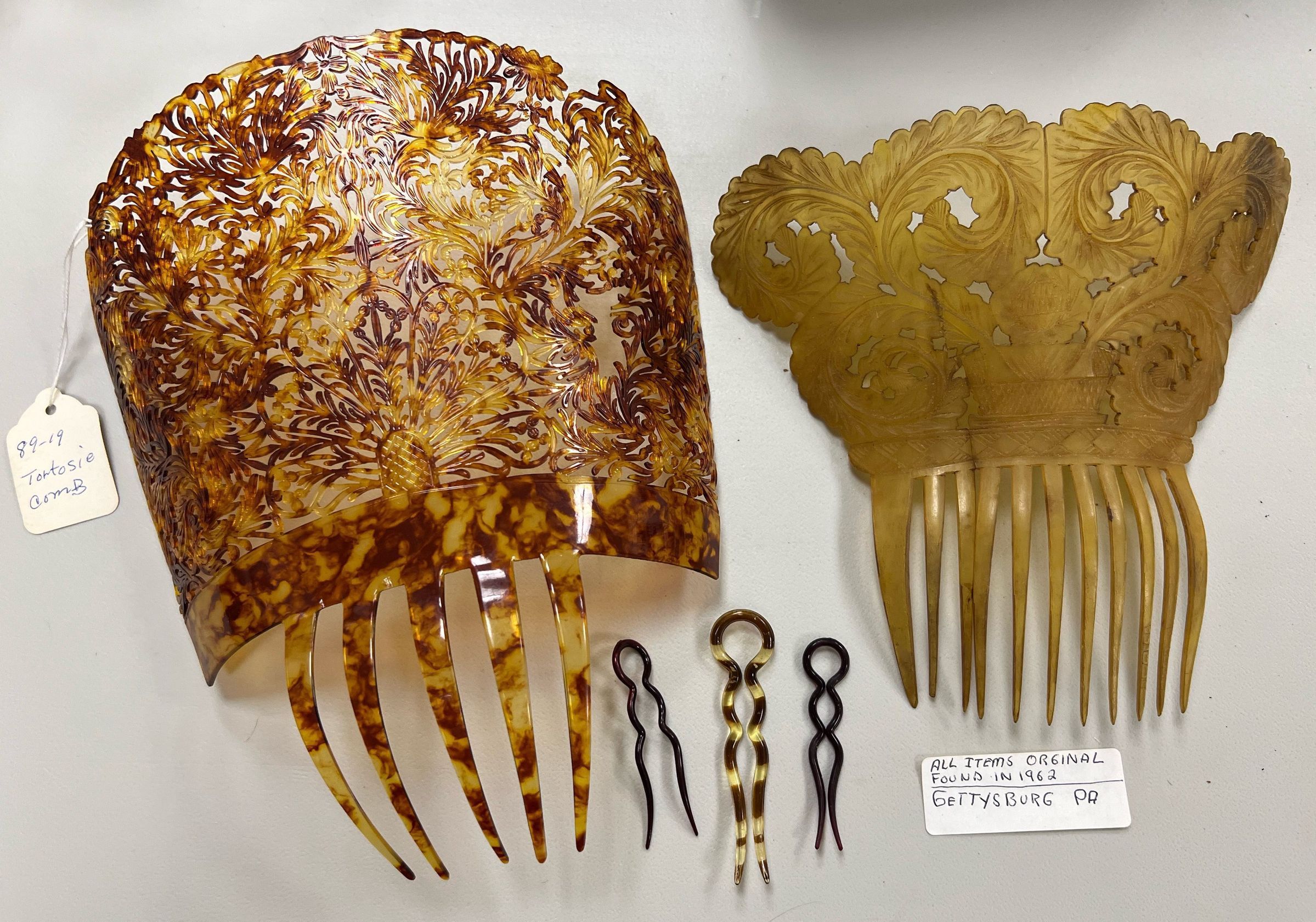The image captures two large ornamental combs alongside a set of smaller hair accessories, all displayed against a white background. On the left, a large tortoiseshell comb features intricate floral patterns, while the right displays a yellowish comb with elegant cut-outs and similarly floral motifs. A tag notes that these items are originals found in 1962 in Gettysburg, Pennsylvania, emphasizing their historical significance.