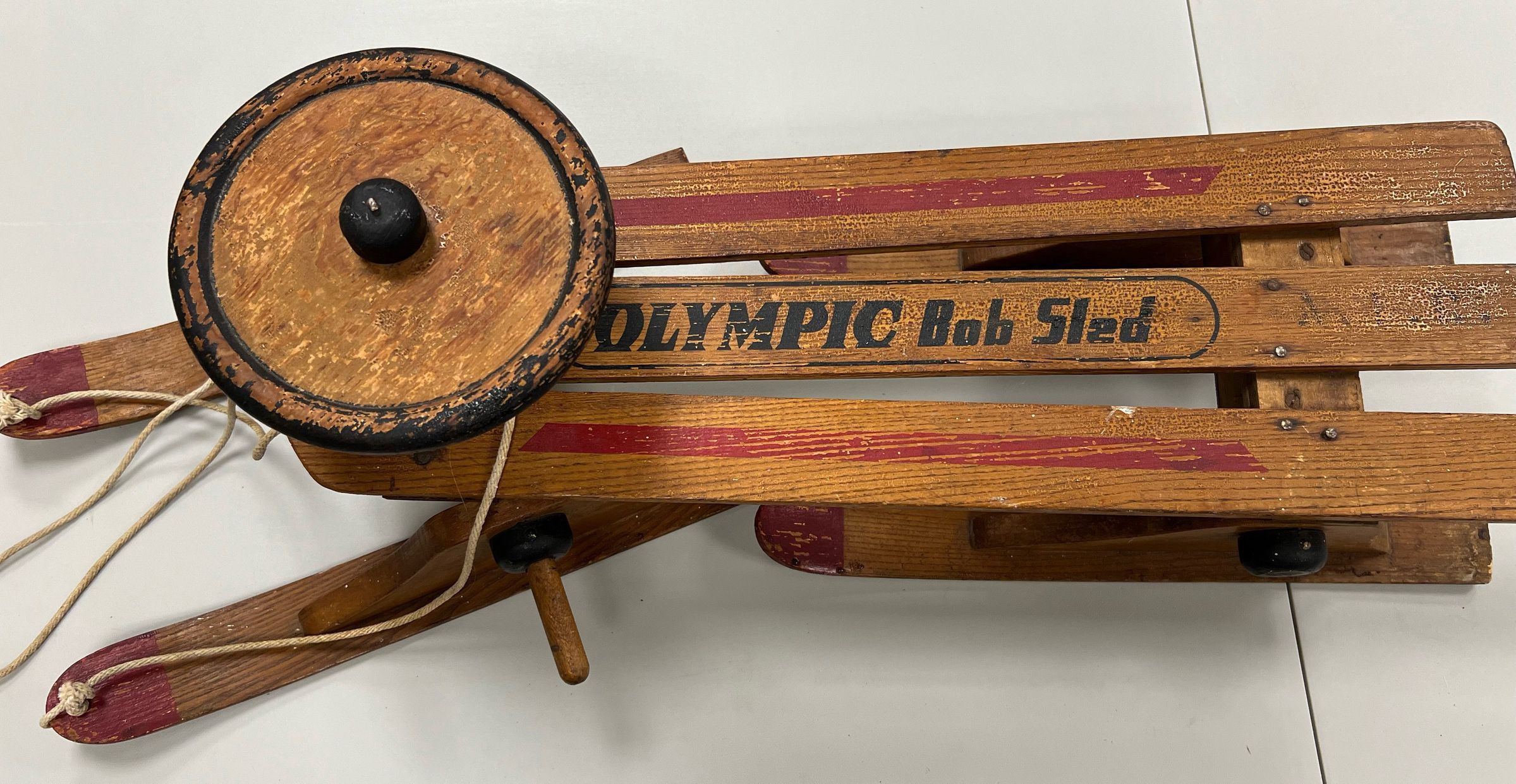 The image shows a vintage wooden sled, characterized by its robust, rustic design, featuring faded red and yellow paint on its horizontal slats. The sled is branded with the words "OLYMPIC Bob Sled," suggesting a theme or homage to Olympic bobsledding, and it includes a steering mechanism topped with a black circular grip, connected by ropes for control. The contrast of the battered wood and worn paint hints at its considerable use and adds to its antique charm, presenting a nostalgic