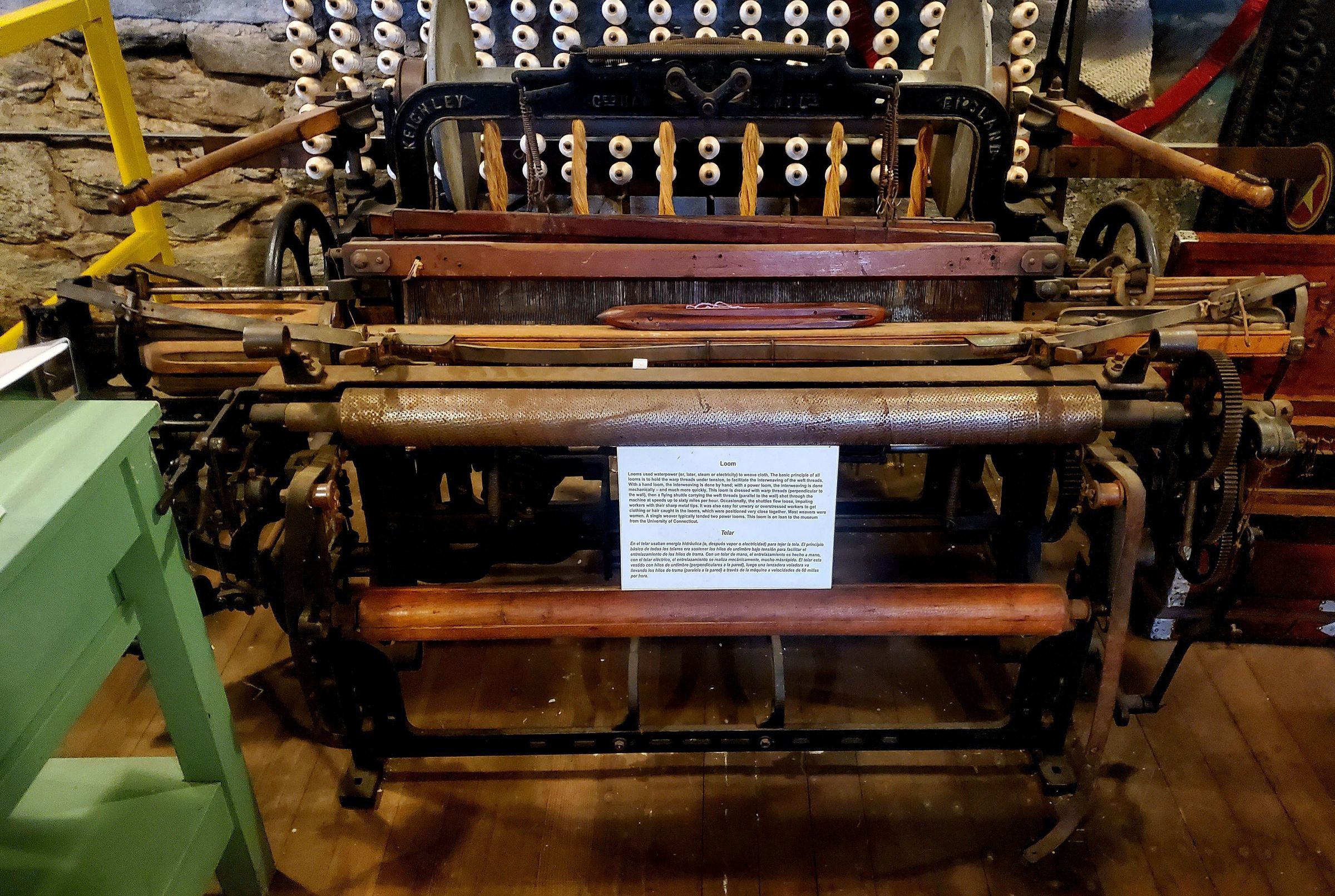 Hattersley Domestic Loom: Hattersley Domestic Loom manufactured by George Hattersley and Sons of Keighley, England. On semi-permanent loan from the University of Connecticut. "The first Hattersley loom was developed by George Hattersley and Sons of Keighley, West Yorkshire, England. The company had been started by Richard Hattersley after 1784, with his son, George Hattersley, later entering the business alongside him. The company developed a number of innovative looms, of which the Hattersley Standard Loom – developed in 1921 – was a great success." (Wikipedia.) "The plain Hattersley Domestic Loom was specially developed for cottage or home use and designed to replace the wooden handloom; the Domestic is similar in construction to a power loom. It was introduced ca.1900 and the makers claimed that a speed of 160 picks per minute could be easily attained with from 2 to 8 shafts weaving a variety of fabrics. Because foot pedals, or treadles, operate the loom it is still classed as a handloom, but it is much easier and faster to weave as all the motions of the loom are connected via crankshaft and gear wheels. The cast metal chair, manufactured along with the loom, can be raised or lowered to suit, and the seat rocks forward and back as the weaver treadles the loom." (Wikipedia.) See also https://www.keighleynews.co.uk/news/11028713.historic-keighley-loom-helps-celebrate-textiles-heritage/.
