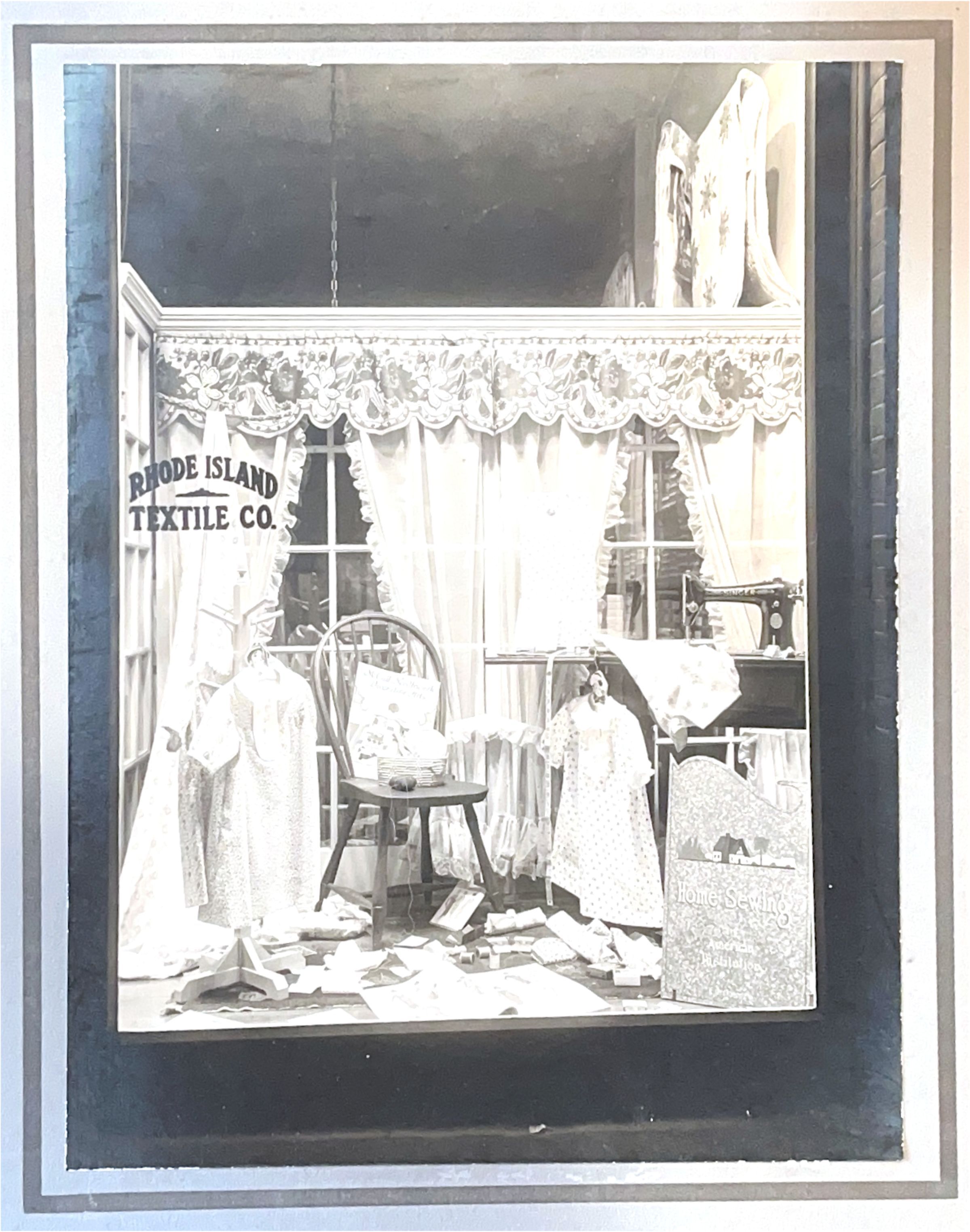 Photograph of Rhode Island Textile Co. Window: The image depicts a vintage shop display from "Rhode Island Textile Co." showcasing a home sewing scene. In the window are various textiles and garments on display, including hanging dresses and folded fabrics, suggesting a focus on domestic sewing projects. A wooden chair holds a stack of pattern booklets, reinforcing the sewing theme. Surrounding the chair, decorative curtains with floral patterns frame the space. The floor is strewn with additional sewing patterns and fabric samples, creating an impression of a bustling and creative work area. The overall presentation emphasizes a warm, homely atmosphere, inviting passersby to explore the joys of home sewing.