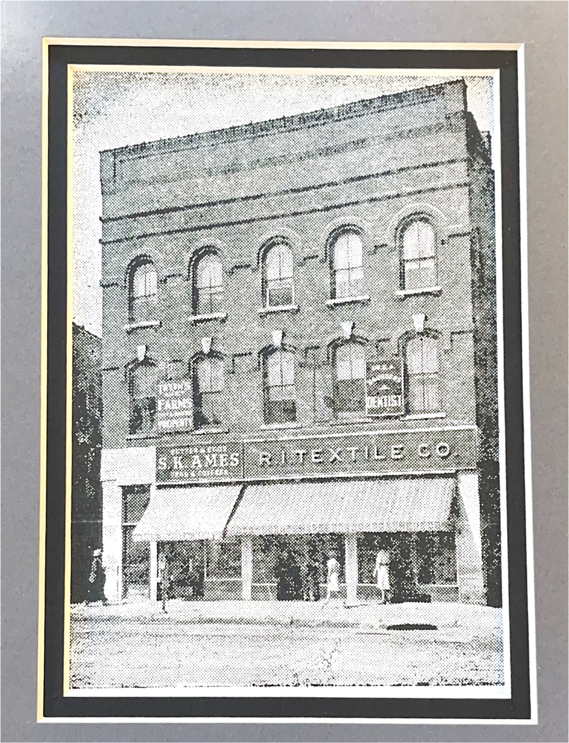 The image displays an old black-and-white photograph of a three-story brick building, featuring arched windows on the upper floors and an awning over the entrance at street level. The signage on the facade reads "S.K. AMES" and "R.I. TEXTILE CO.," suggesting a textile company. Additional signage indicates a dentist's office and a farm-related business, highlighting the multifaceted use of the building.