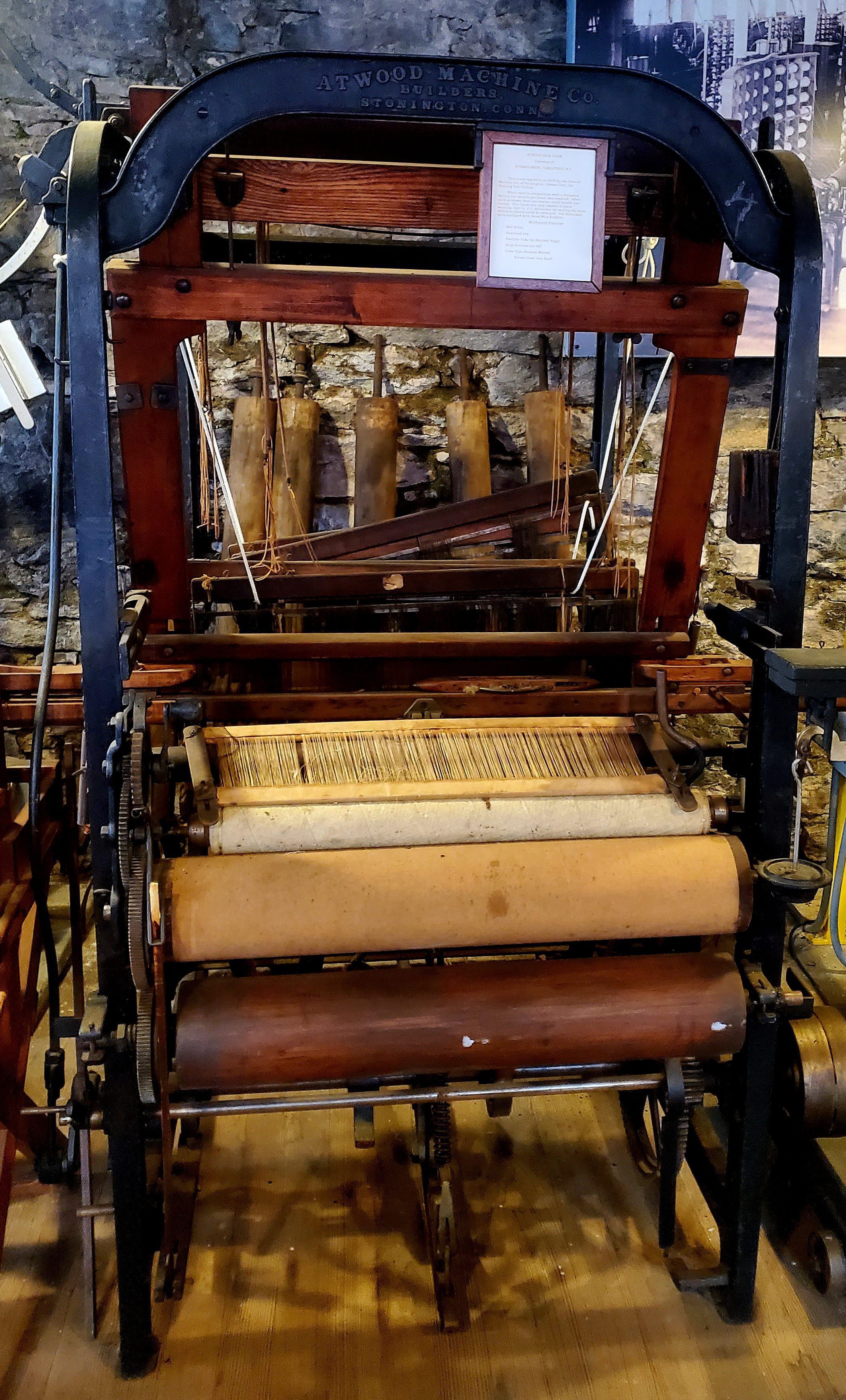 Atwood Industrial Loom: Atwood industrial silk loom. This loom was built in 1875 by the Atwood Machine Co. of Stonington, CT, for weaving silk textiles. When used in conjunction with a Jacquard Motion one weaver per loom was required; when used as shown here one weaver could handle two looms. This loom was only capable of plain weaving; that is, 1/1 fabrics but by varying the draw different effects could be obtained. The harnesses were equipped with Swiss wire heddles. Belt drive. Overhead lay. Positive take-up (ratchet type). Rope friction let-off. Cone type harness motion. Driven from cam shaft. The Atwood Company began in Mansfield, CT, relocated to Willimantic, CT, and then relocated again to Stonington just before this loom was constructed. The Mill Museum acquired it from the Little Red Shop Museum in Hopedale, MA. The LRSM is located in a wooden building that was once the original building of the Draper Company, once the United States' major manufacturer of industrial looms. As the Draper Company grew, it moved its manufacturing to larger brick buildings and converted the original building into a museum that featured a variety of looms, some made by Draper and some not, from different eras, including this Atwood loom. When the Draper Company closed, the museum became the property of the Town of Hopedale and today is operated as a local history museum. Not having a need for looms made outside of Hopedale, the LRSM donated this loom to the Mill Museum.