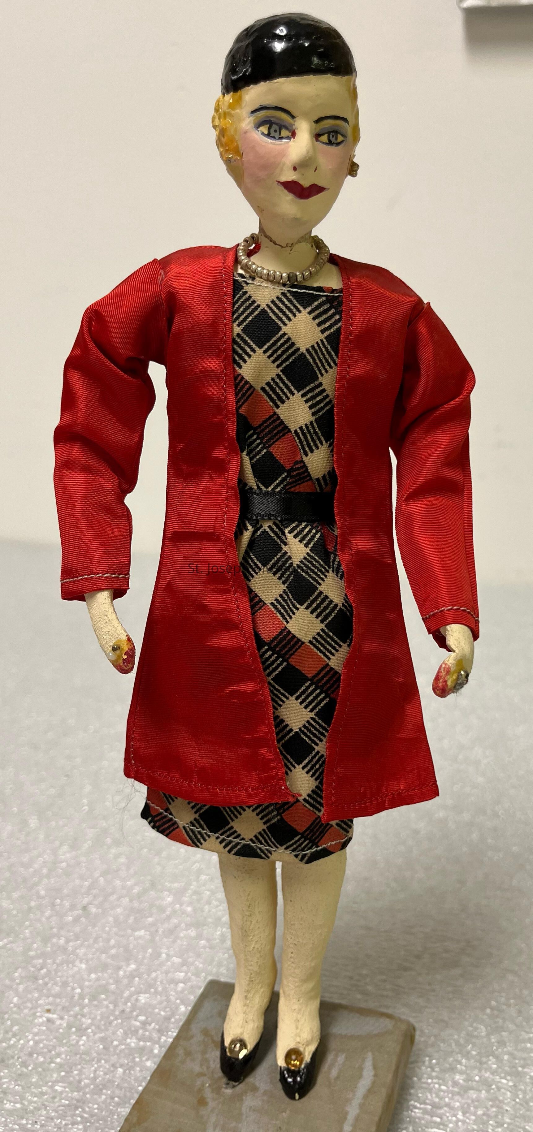 The image shows a handcrafted doll with distinct, stylized features, wearing a black and beige plaid dress underneath a vibrant red jacket. The doll has a short, black hairstyle and painted facial features, including blue eyes and red lips. Around its neck is a beaded necklace, adding to its detailed appearance. The hands, painted with red nails, are positioned at the sides, while the feet are adorned with black shoes featuring gold embellishments. The doll stands upright on a simple rectangular base, with the texture of its surface visible, suggesting it might be handmade. This figure embodies a vintage or classic style, highlighted by its choice of clothing and careful detailing. The overall craftsmanship and color contrast between the outfit and the doll’s features make it visually striking and indicative of traditional doll-making techniques, likely intended for decorative purposes rather than play.