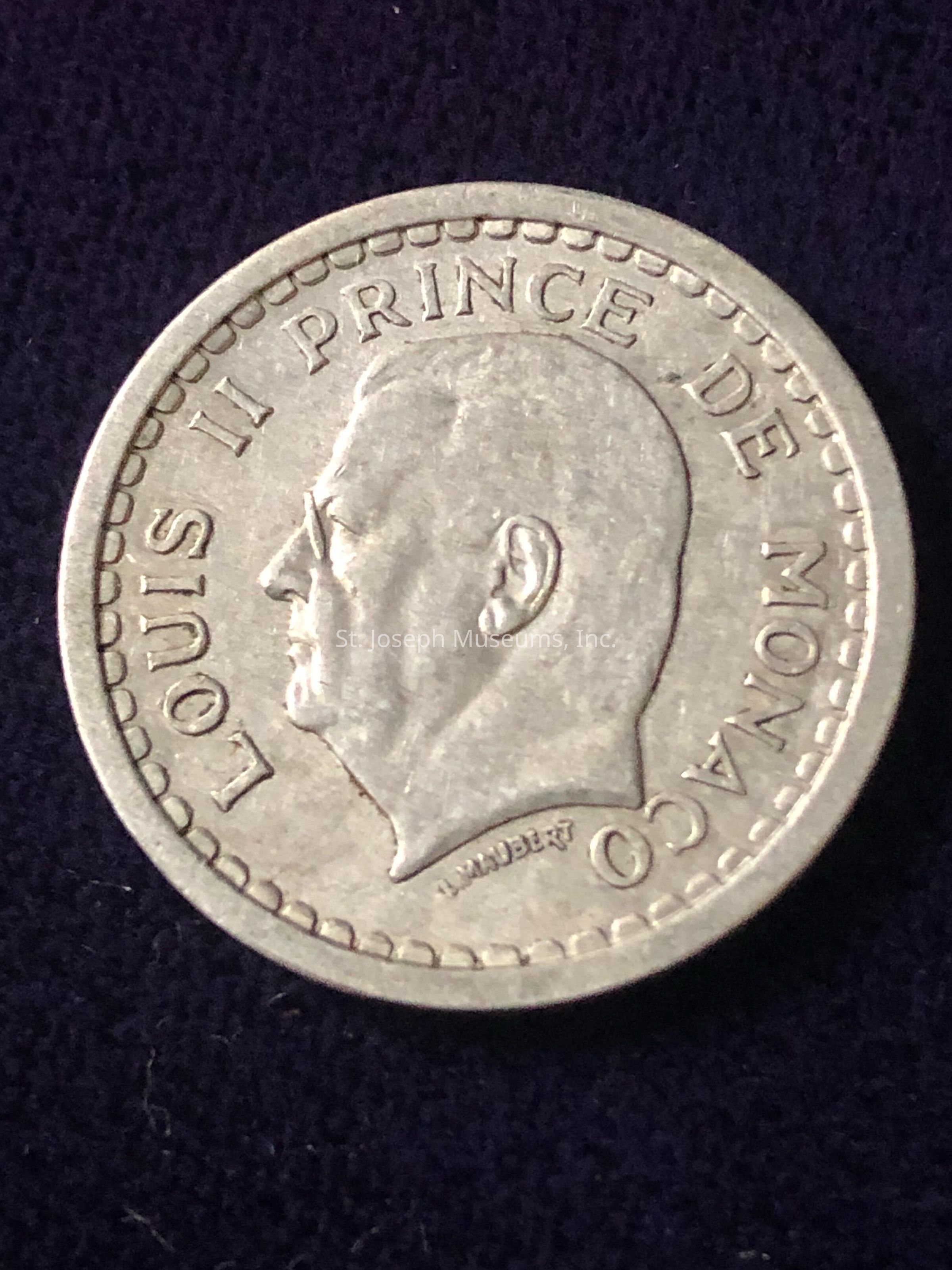The image displays a coin featuring a profile portrait of a person in relief, surrounded by the inscription "LOUIS II PRINCE DE MONACO." The coin has a beaded border along its edge, giving it a classic appearance. Additionally, there’s a small inscription near the neck of the portrait that reads "LAMARRE." The design suggests it commemorates Louis II, a historical figure linked to Monaco. The coin appears to be struck in a metallic, likely silver or a silvery alloy, and rests against a dark background, enhancing its shiny surface and intricate detailing.