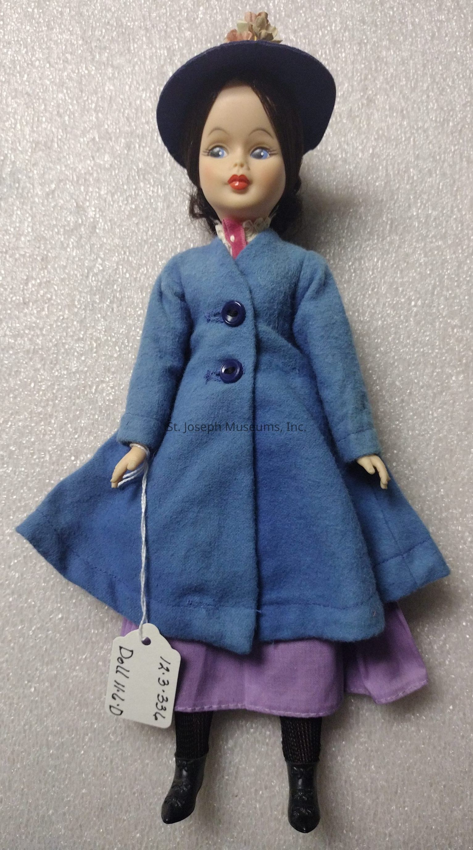The image depicts a vintage-style doll set against a textured, light background. The doll is dressed in a vibrant ensemble featuring a long, light blue coat with two buttons down the front, layered over a purple dress. She wears a stylish blue hat adorned with small flowers on top. Her facial features include painted blue eyes and red lips, giving her a classic, elegant look. The doll's legs are covered with dark socks, and she is wearing black shoes. Attached to the doll’s left hand by a string is a tag with inscriptions that include "1.1.3.334" and "Doll No. 10," providing details likely for identification or cataloging purposes.