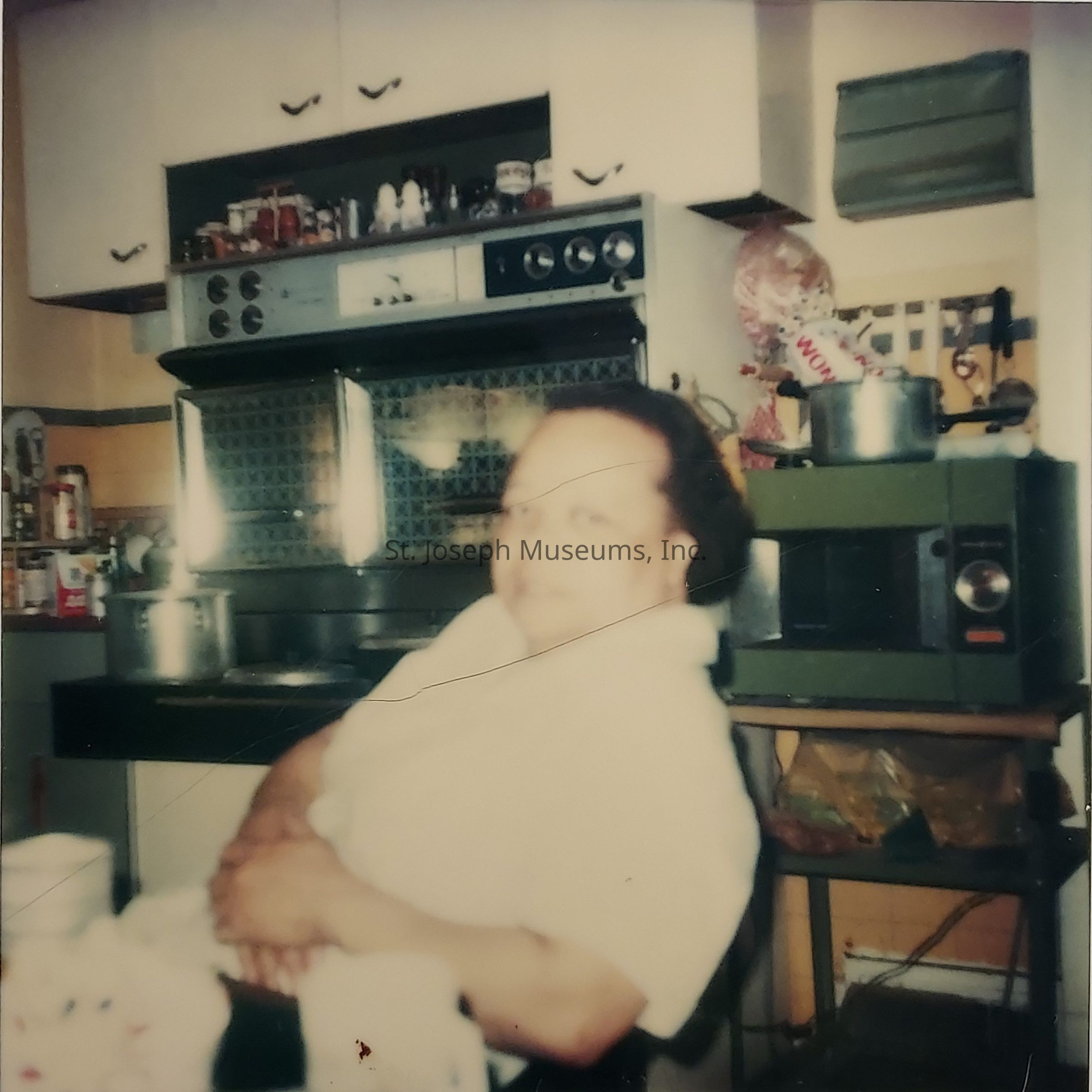 In this vintage-style photograph, a person is seated in a cozy kitchen, wearing a light-colored blouse. The kitchen is equipped with an older model oven and a microwave positioned against the wall. Several pots and kitchen items are placed on top of the appliances, giving a warm and lived-in feel. Above the oven are spice jars and condiments, indicating a space well-used for cooking and meal preparation. The overall ambiance of the kitchen suggests a homely and nostalgic setting, with warm lighting that enhances the retro atmosphere. The colors are somewhat faded, adding to the vintage charm of the image.