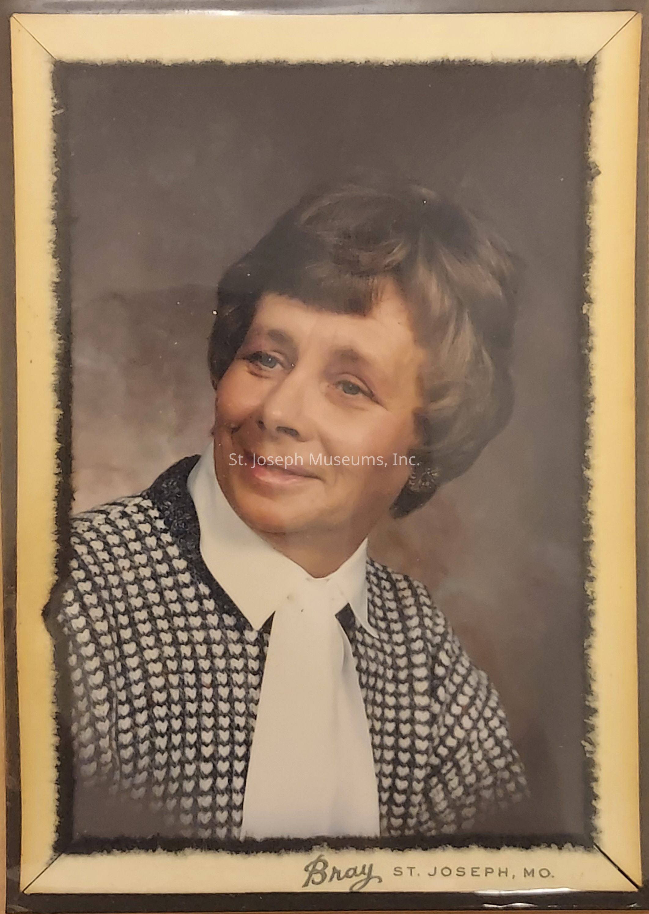 The image is a framed portrait of an older woman with short, styled hair, wearing a textured black and white checkered jacket over a white blouse with a high collar and a bow, and she gazes slightly to her right with a gentle, composed expression, smiling subtly; the background appears to be a softly blurred, warm-toned backdrop, and the frame has a classic style with a beige border and an inner black edge, displaying the photographer's mark, "Bray, St. Joseph, MO," at the bottom.