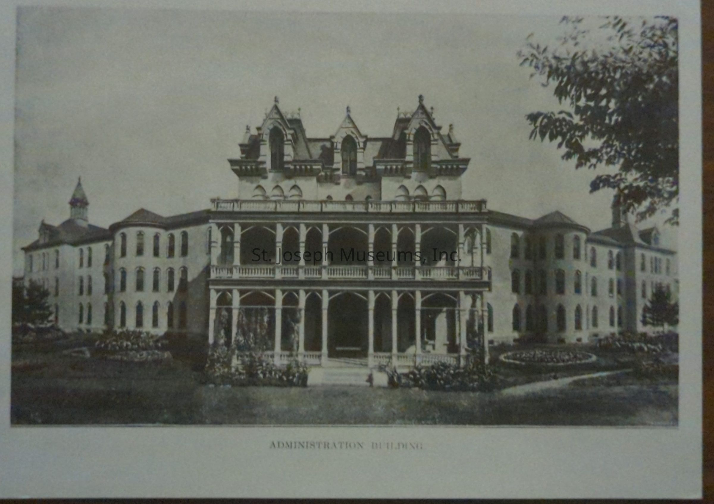Black-and-white photograph of the Administration Building of the State Hospital in St. Joseph, Missouri, located at 36th and Frederick Avenue, is an impressive historic structure featuring elaborate architectural details, including ornate gables, two-story balconies, and a grand entrance, framed by meticulously maintained gardens and mature trees.