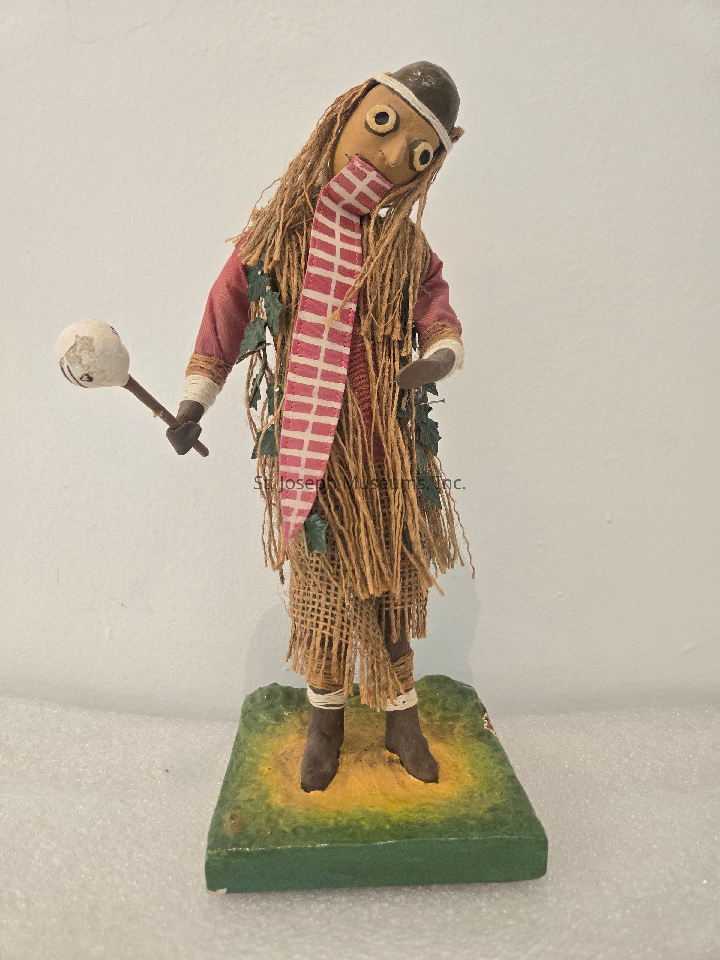 The image features a whimsical handcrafted figurine standing on a green square base, adorned with a wide-eyed expression and a playful demeanor. It wears a red shirt with the sleeves and a light brown skirt woven with burlap-like material, with leafy accents adding a natural touch. The figure's head is topped with a brown rounded cap, and it has long, textured hair cascading down made of fibers resembling straw or yarn. A pink and white checkered scarf is wrapped around its neck and falls down the front, enhancing its quirky appearance. In one hand, it holds a staff, topped with a light-colored sphere, with the hand appearing open. The overall scene exudes an imaginative, folkloric charm, blending elements of whimsy and craftsmanship, likely intended to conjure visions of a rustic character or festive figure deeply rooted in cultural storytelling or artistic expression. The background is plain and white, drawing full attention to the intricate details and textures of the figure itself.