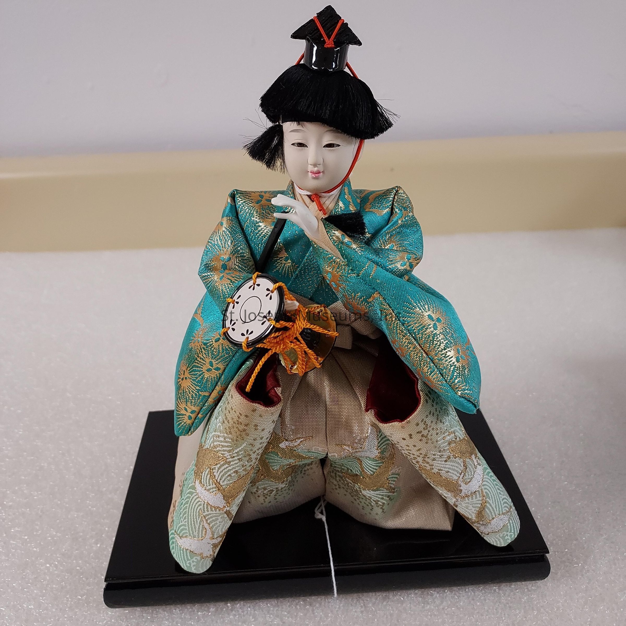 The image shows a traditional Japanese doll seated on a black rectangular base, adorned in an elaborate teal and gold kimono featuring intricate gold patterns. The doll has a delicately painted face with fine black eyebrows and closed eyes that exude a serene expression. Mounted on its head is an ornate black headdress tied with red strings, adding to its regal appearance. The doll's hands, meticulously carved and painted, hold a small traditional drum, wrapped in orange strings. The attire's vibrant hues and meticulous detailing highlight the craftsmanship and cultural significance of the doll, reflecting Japanese artistic traditions.