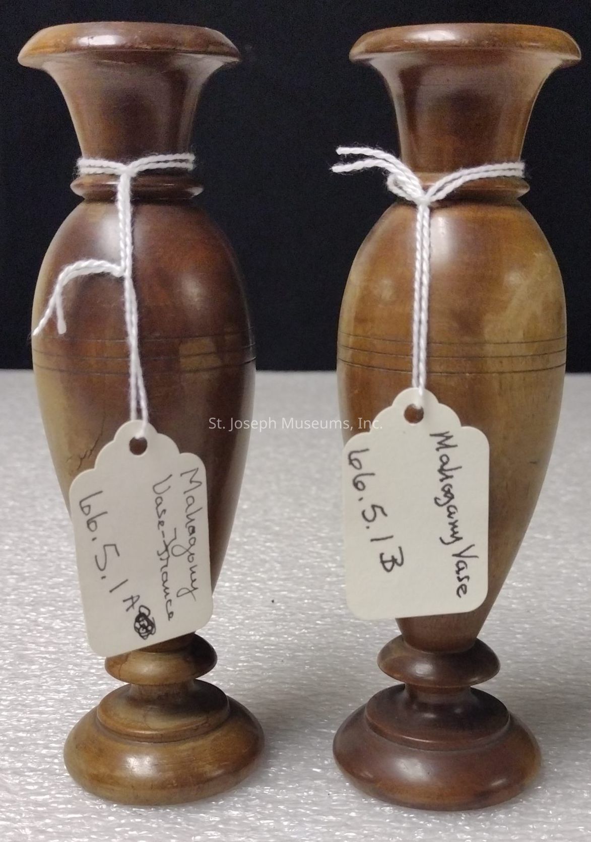 The image shows two elegant, small mahogany vases with a polished finish, each standing on a round pedestal base, and featuring wide, flared rims. These vases are tied with white string, from which tags hang. The tags indicate their classification as "Mahogany Vase" and they have identification numbers written on them: 66.5.1 A France and 66.5.1 B, suggesting they might be part of a collection or inventory. The rich, warm brown tones of the vases are highlighted under the lighting, and they rest on a textured, light surface, with a dark background accentuating their presence, making them appear as finely crafted decorative pieces that likely hold historical or artistic significance.