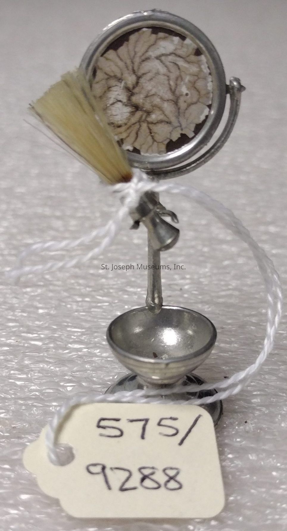 The image depicts a miniature silver-colored microscope-like apparatus with a small brush attached to the side, which stands on a textured surface. The apparatus features a circular frame holding what appears to be a thin, dried anatomical specimen that resembles a brain or intricate leaf pattern. At the base of the structure is a tiny, shallow bowl or dish, which could serve as a stand or a container for small items. A white string is looped through a hole in the base, connecting it to a cream-colored tag inscribed with the numbers "575/9288" in black ink, suggesting cataloging or inventory identification. The entire setup adds a sense of curiosity, possibly used for scientific demonstration or as a collectible.
