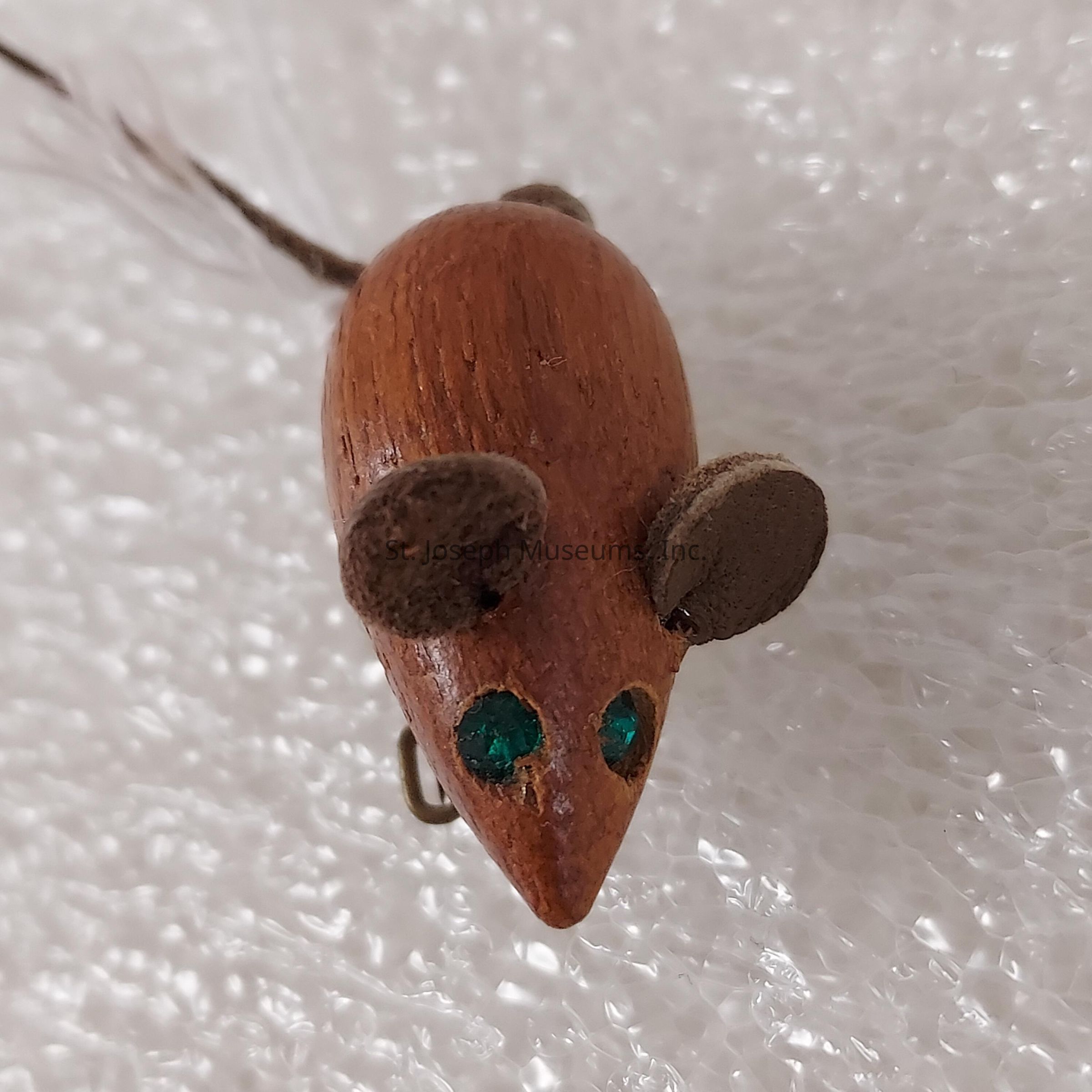 The image depicts a small, intricately carved wooden object resembling a mouse, complete with detailed features such as tiny, round ears and glimmering green gemstone-like embellishments for eyes. Its smooth, polished texture gives it a lifelike appearance, while the subtle wood grain adds depth and character to its surface. Attached to the rear is a slender tail that gracefully curves, enhancing the overall realism of the miniature creation. The mouse sits on a textured white background, which contrasts with its warm, natural colors, allowing its craftsmanship to be the focal point of the scene, showcasing the artisan's attention to detail.