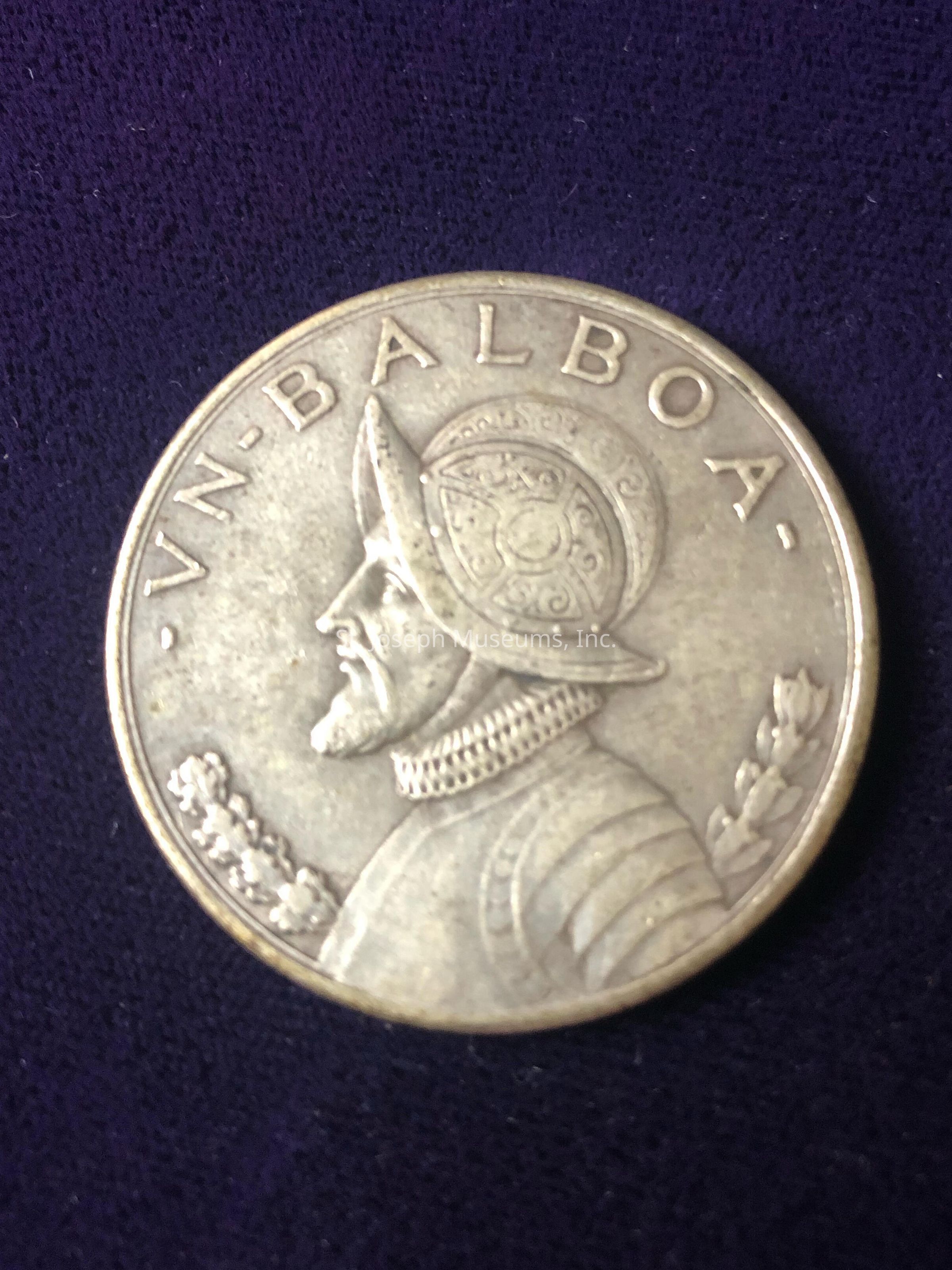 The coin is inscribed with "VN BALBOA" at the top, and it prominently displays the profile of an armored figure wearing a detailed helmet. The figure's armor and helmet are intricately designed, featuring decorative patterns, while laurel branches are depicted on either side of the figure, emphasizing the theme of honor and achievement. The coin appears metallic, possibly silver or a similar material, and exhibits a worn yet well-preserved quality, suggesting it might be of historical significance or collectible value.