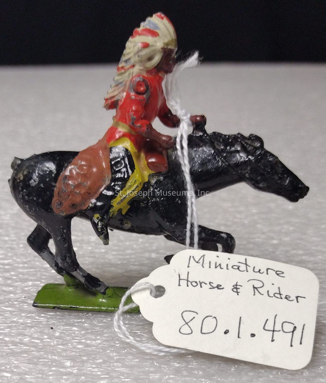 The image depicts a small figurine of a rider atop a horse. The rider is adorned in a red outfit with a detailed headdress, suggesting a theme of native cultural attire. The horse is painted black and stands on a green rectangular base. Attached to the figurine is a white tag with the handwritten text "Miniature Horse & Rider" followed by the numbers "80.1.491". The craftsmanship appears focused on capturing the dynamic pose of the rider and horse, making it a collectible or decorative piece. The background features a neutral, textured surface.