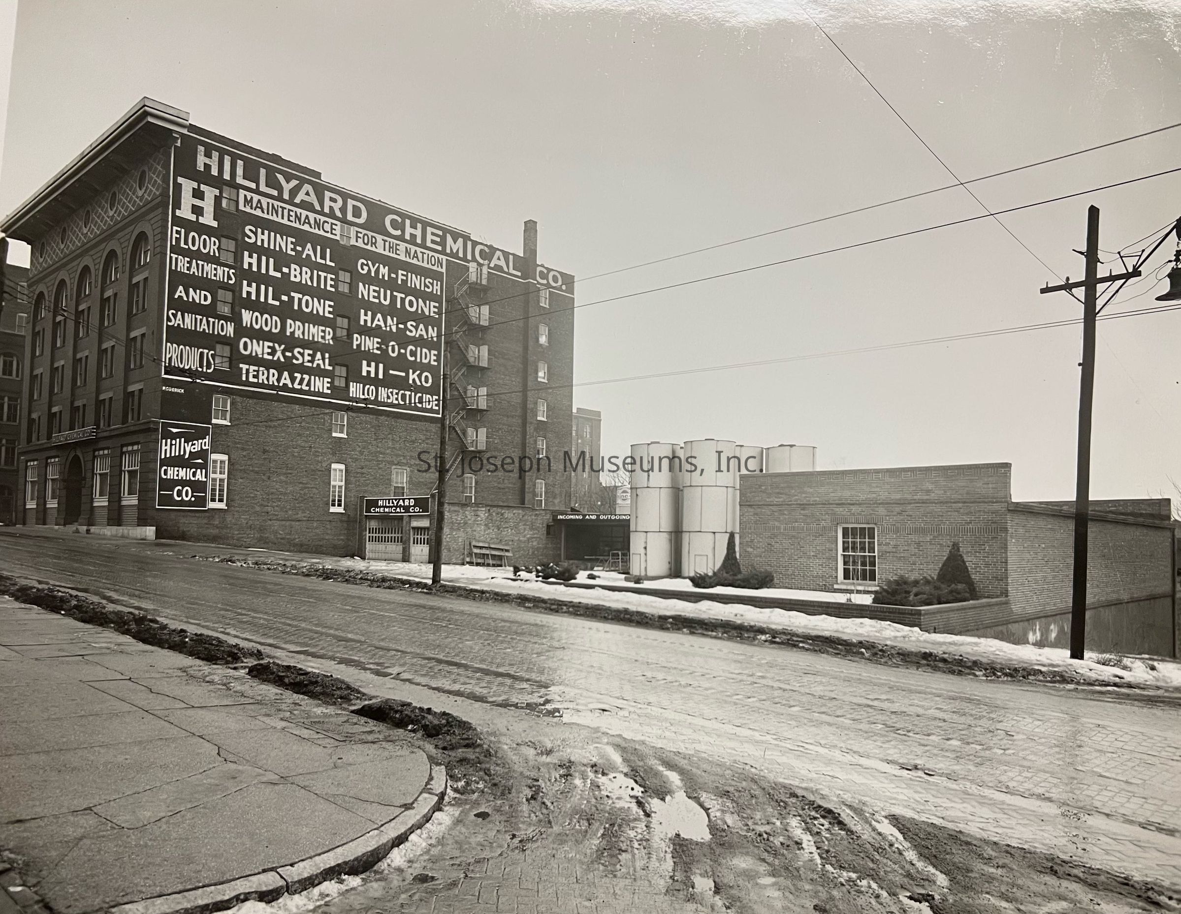 Black-and-white photograph depicting the exterior of the Hillyard Chemical Co. building, featuring a large sign advertising their various products like floor treatments, sanitation items, and more. The structure is situated at a corner, showing a mix of brick and industrial elements, with tanks and a visible street foreground.