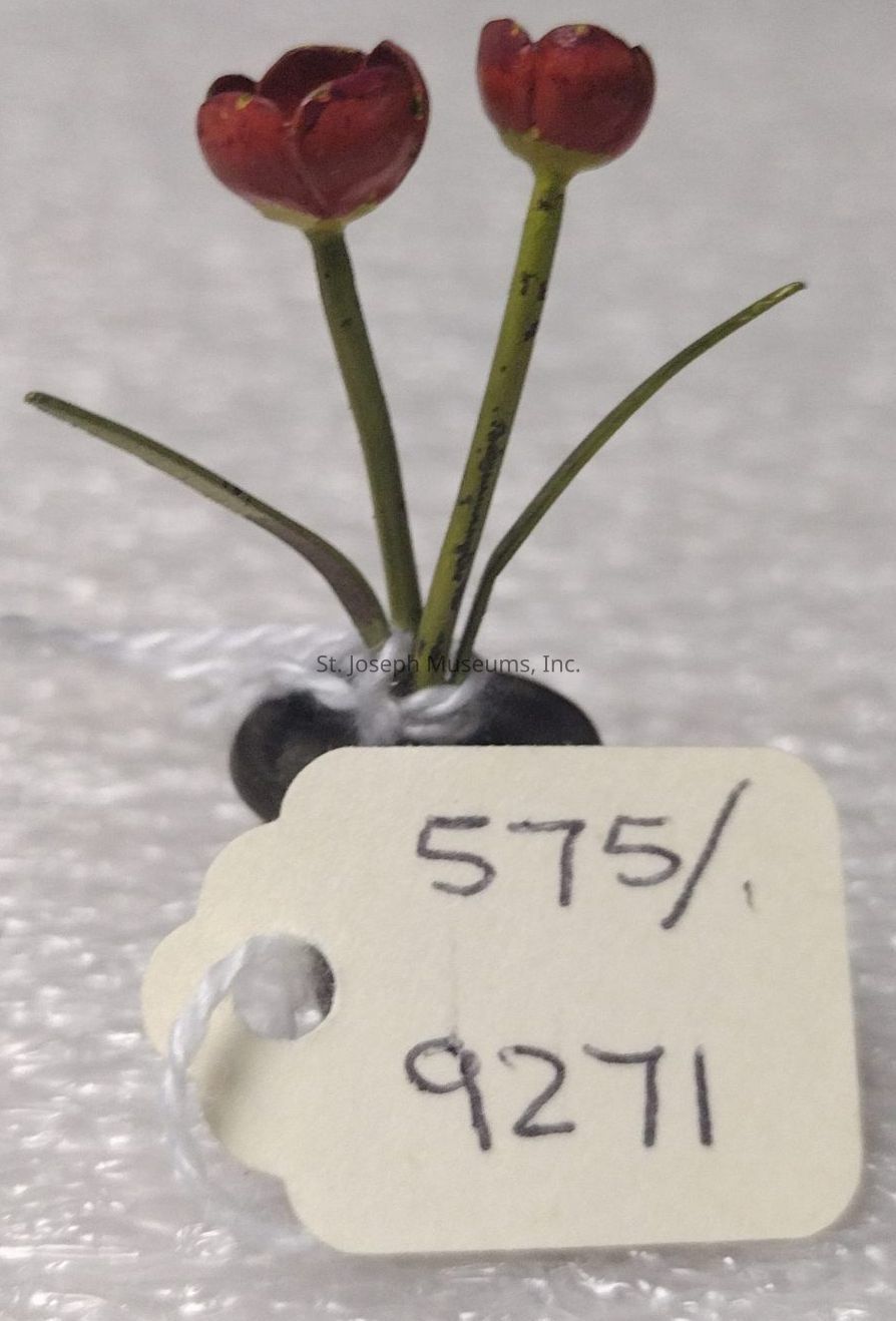 The image shows a small decorative item featuring two red tulip-like flowers with green stems set in a minimalistic black base. The flowers exhibit a simple, charming design with smooth petals and slender, curved leaves. Attached to the base is a white string that holds a small, rectangular tag bearing the handwritten numbers "575/9271" in black ink. This set is placed on a textured surface, possibly a table or fabric, providing a subtle contrast to the intricate details of the miniature floral arrangement. The lighting is soft, highlighting the delicate craftsmanship and vivid colors of the flowers.